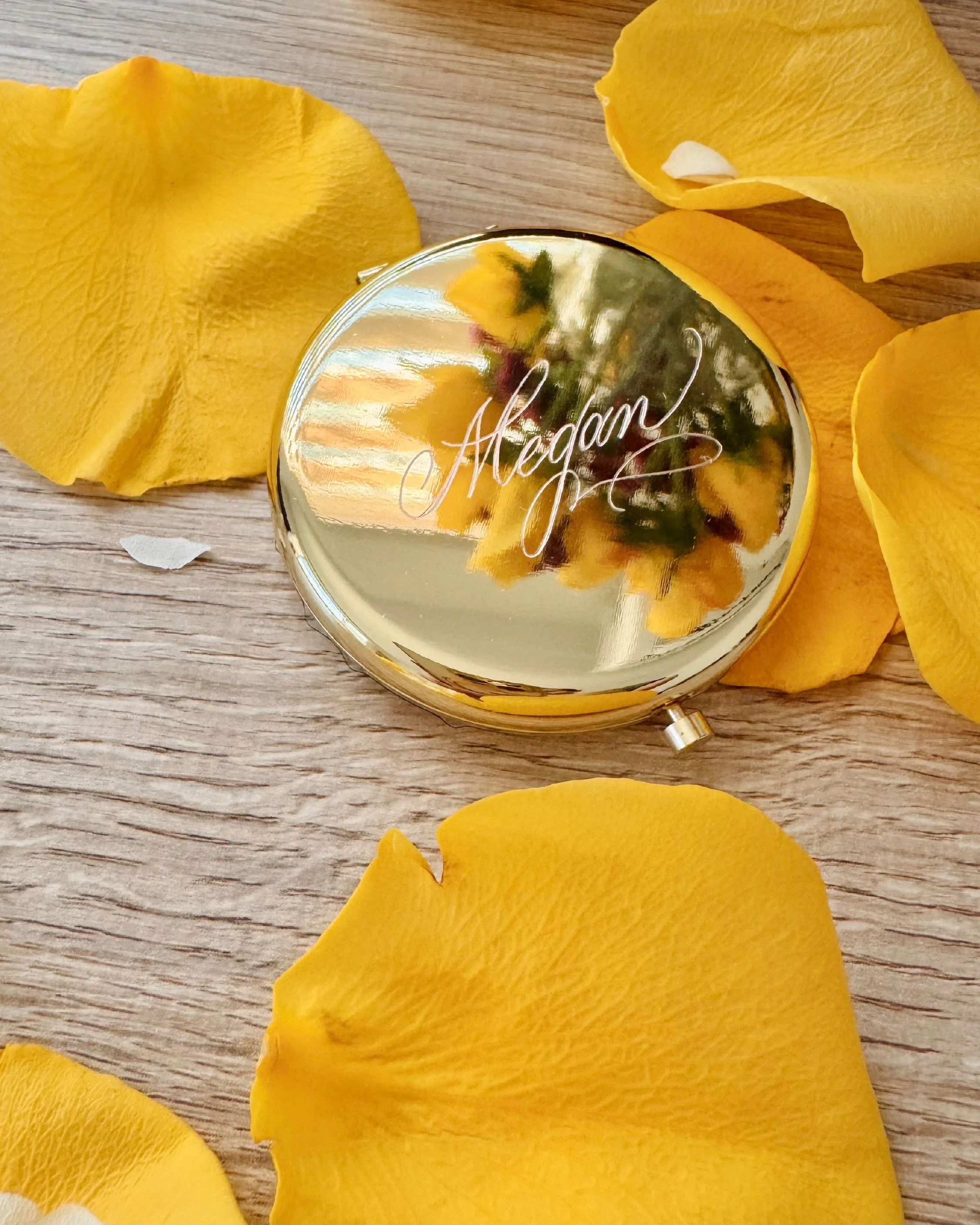 A small, round, gold-colored mirror with the word 'Aegon' engraved on it, surrounded by yellow flower petals, on a wooden surface.