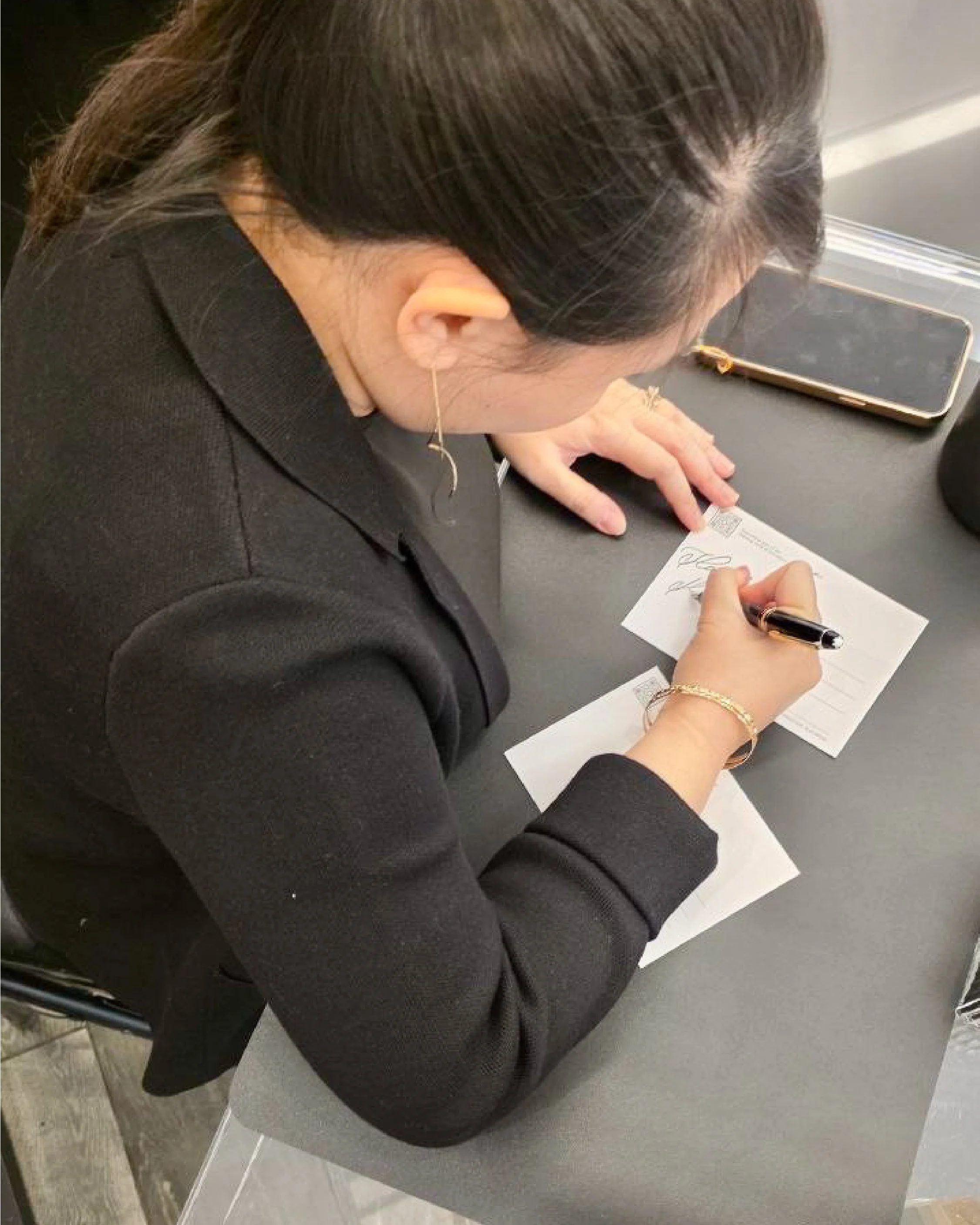 A woman with dark brown hair tied into a ponytail, wearing a black blazer, sitting at a table, signing a document with a black pen, near a smartphone and a black object, on a gray table.