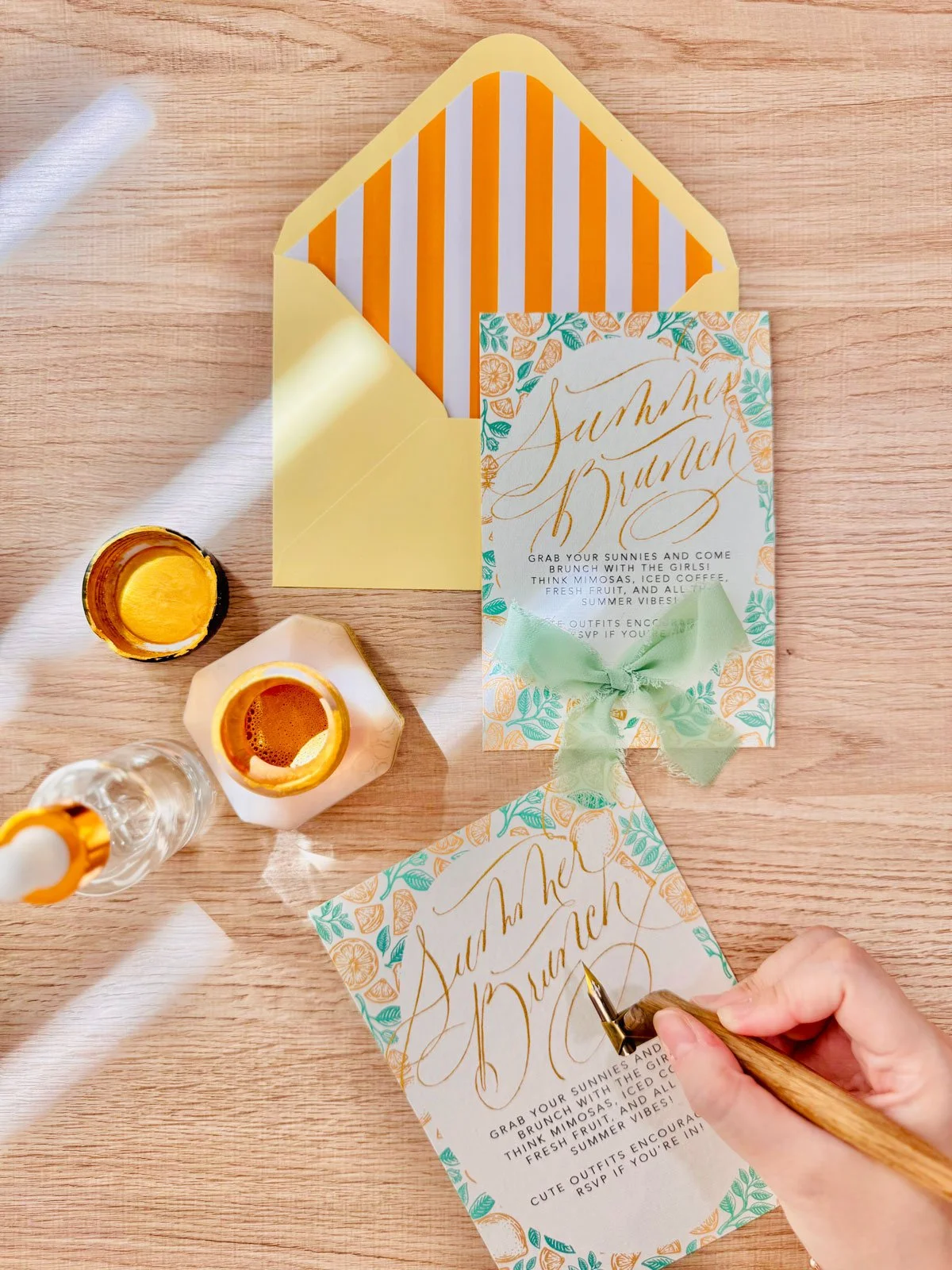Person writing event invitations with gold calligraphy on floral-themed paper, surrounded by yellow and striped envelopes, candles, and drink bottles on a wooden table.