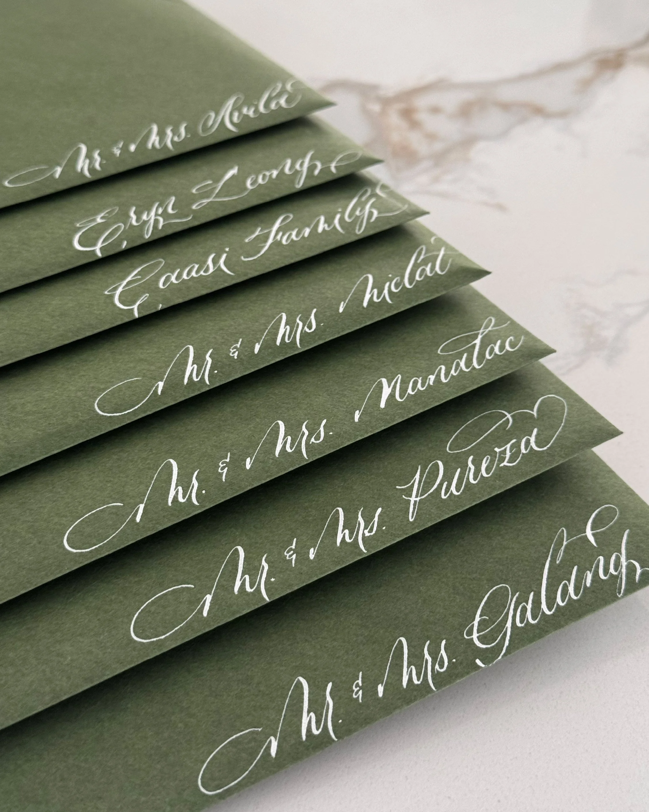 Close-up of layered olive-green cards with elegant white cursive calligraphy, featuring names of couples, arranged on a marble surface.
