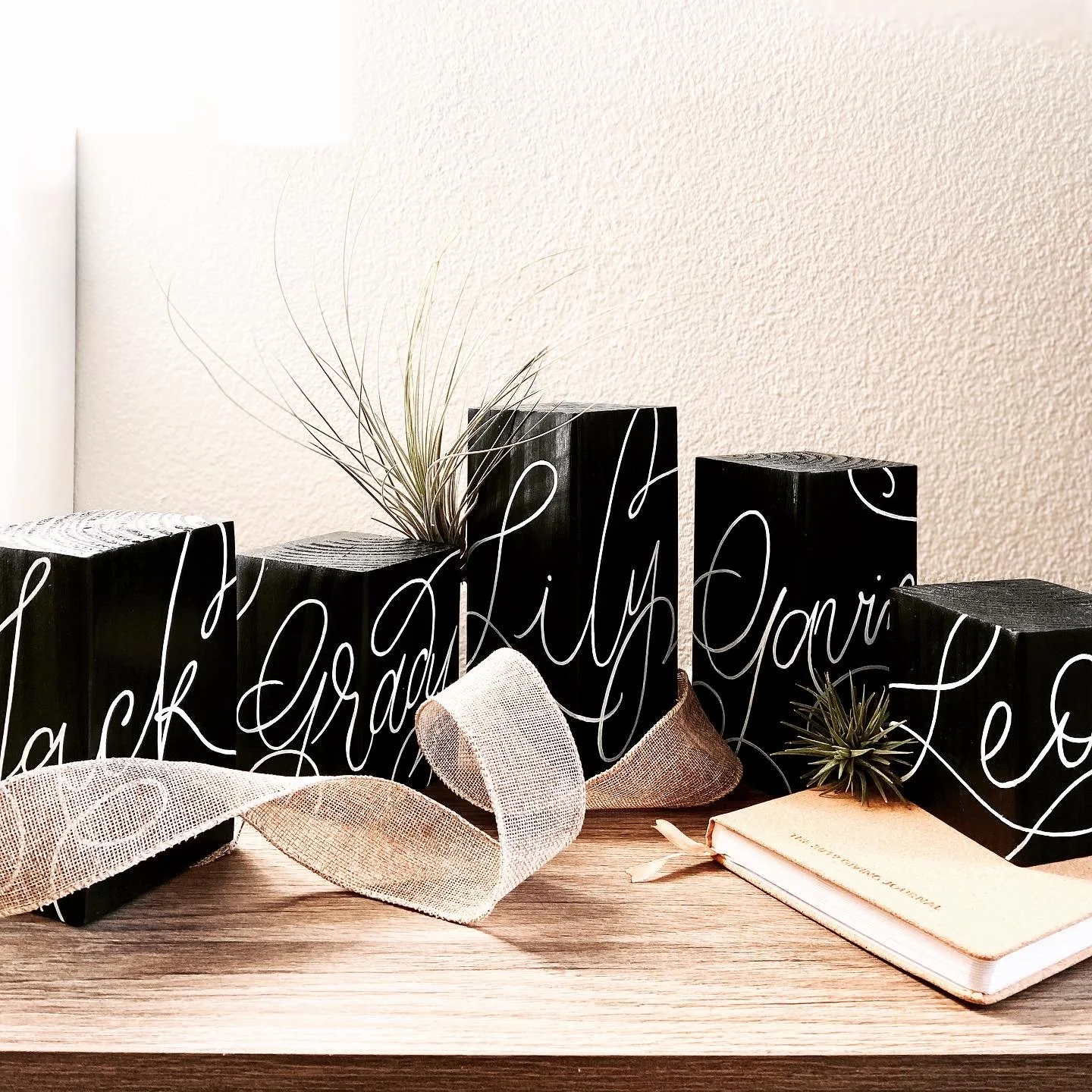 Decorative black blocks with white cursive writing spelling out 'Thankful' and 'Grateful,' with small air plants, a cream-colored ribbon, and a closed journal arranged on a wooden surface against a light beige wall.