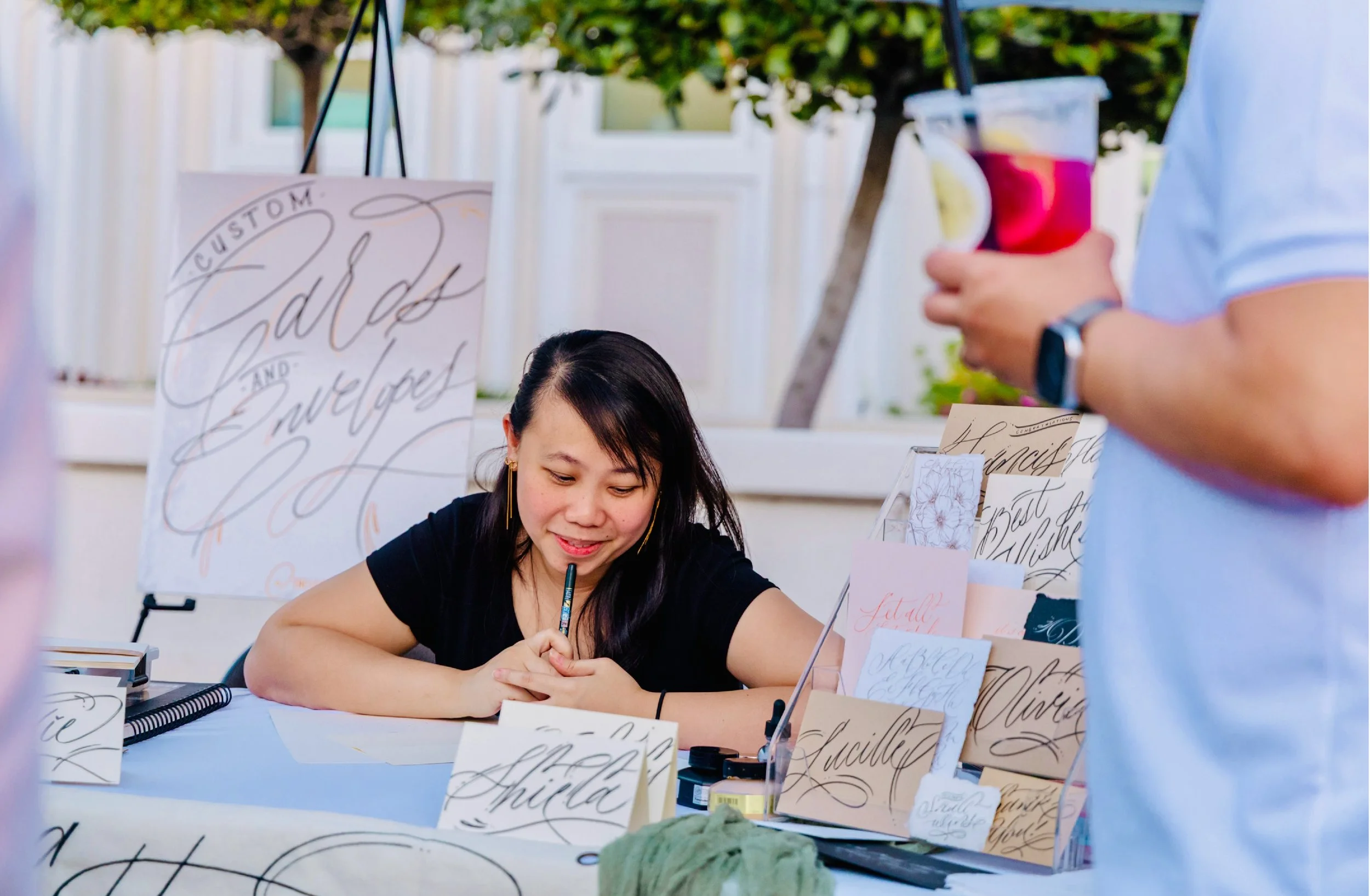 A woman is sitting at a table, signing autographs or artwork with a pen. The table is filled with cards or prints featuring elegant script writing. In the background, there are additional printed materials and a person holding a drink with a slice of lemon.