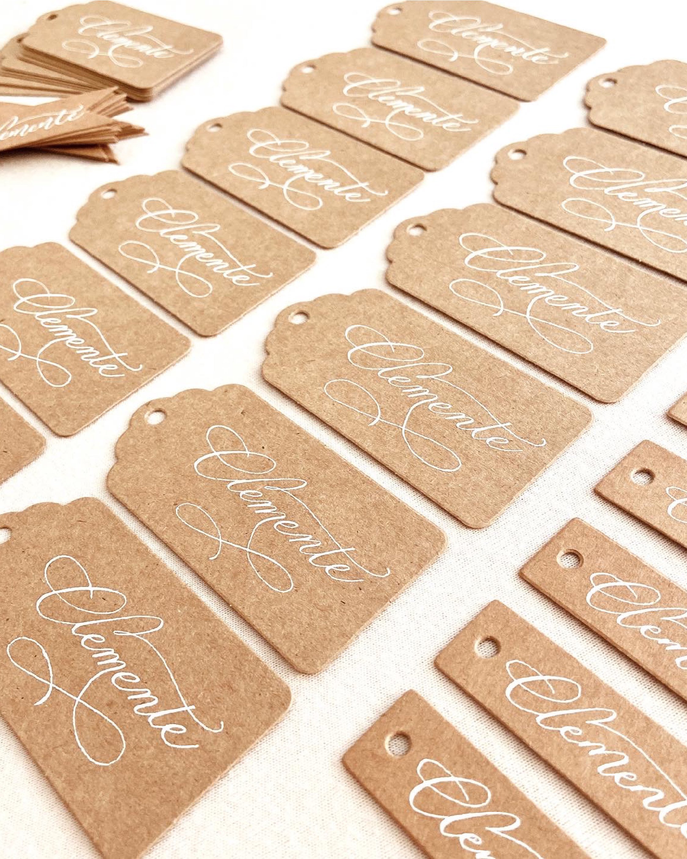 Multiple gift tags made of brown craft paper with white handwritten-style text reading 'Absolutely Love' and a small hole at the top for attaching to gifts. Some tags are stacked, others are arranged in rows on a white surface.