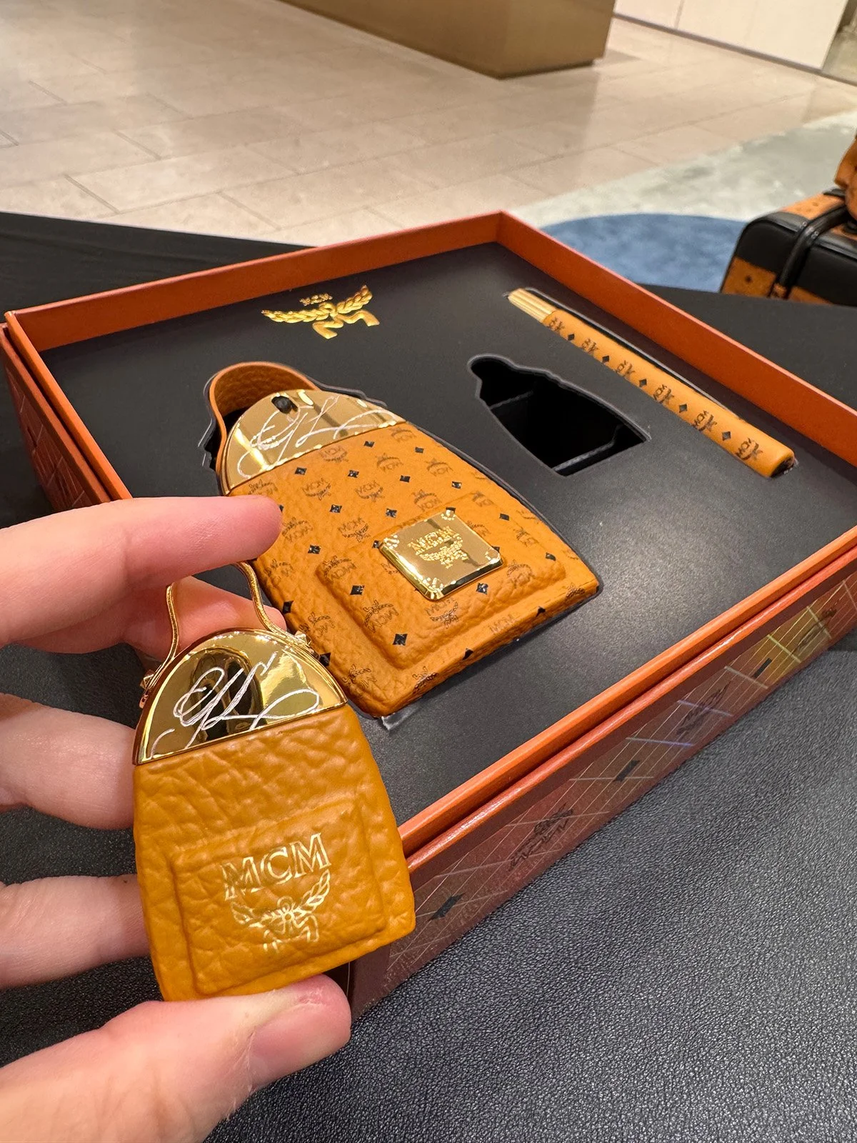 A set of luxurious accessories including a gold high heel shoe, a matching gold and orange bag, a gold case, and an orange keychain all with gold and black accents and branding, placed inside a black and orange box.