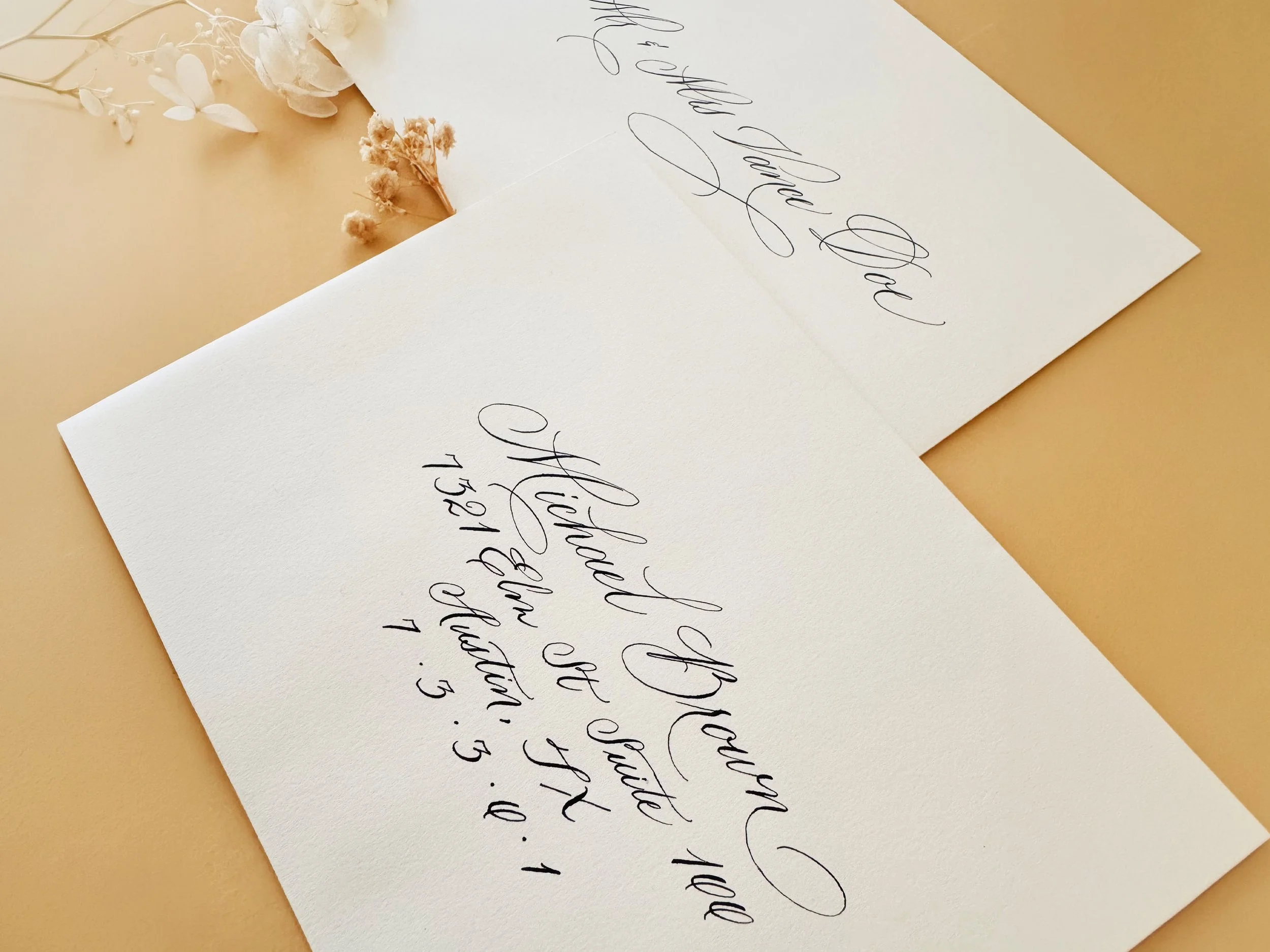 Close-up of a handwritten note with elegant cursive writing, partially covered by dried white flowers on a beige background.