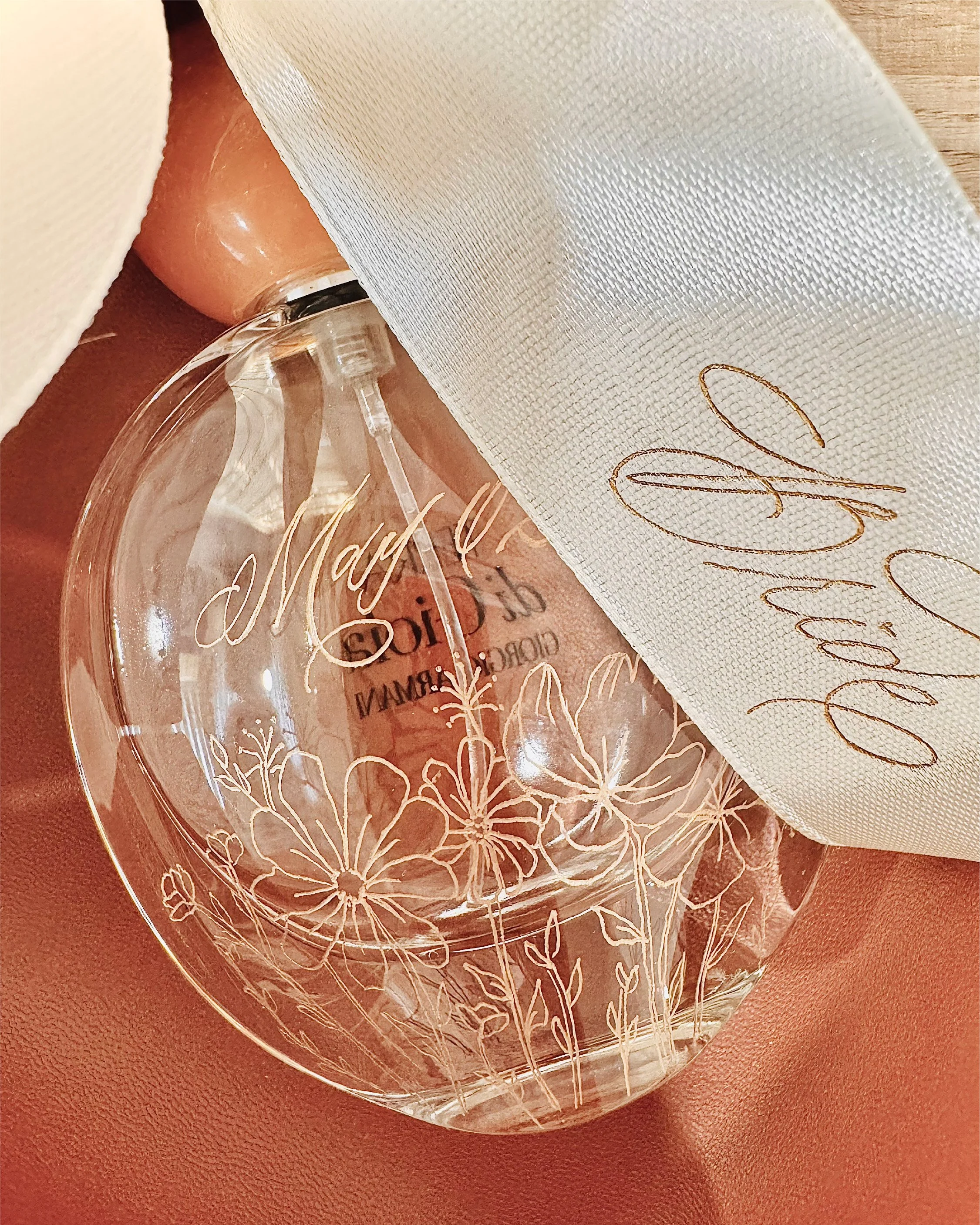 Close-up of a glass perfume bottle with floral engravings, placed on a brown surface with a white cloth covering part of the bottle.