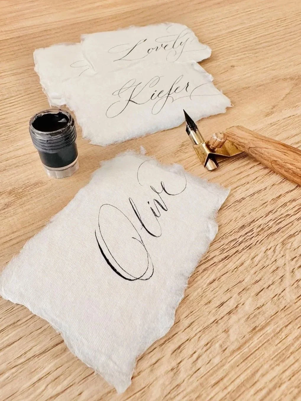 Calligraphy supplies on a wooden table, including a bottle of black ink, a dip pen, and torn pieces of paper with handwritten words 'Lovely' and 'Olive' in elegant script.