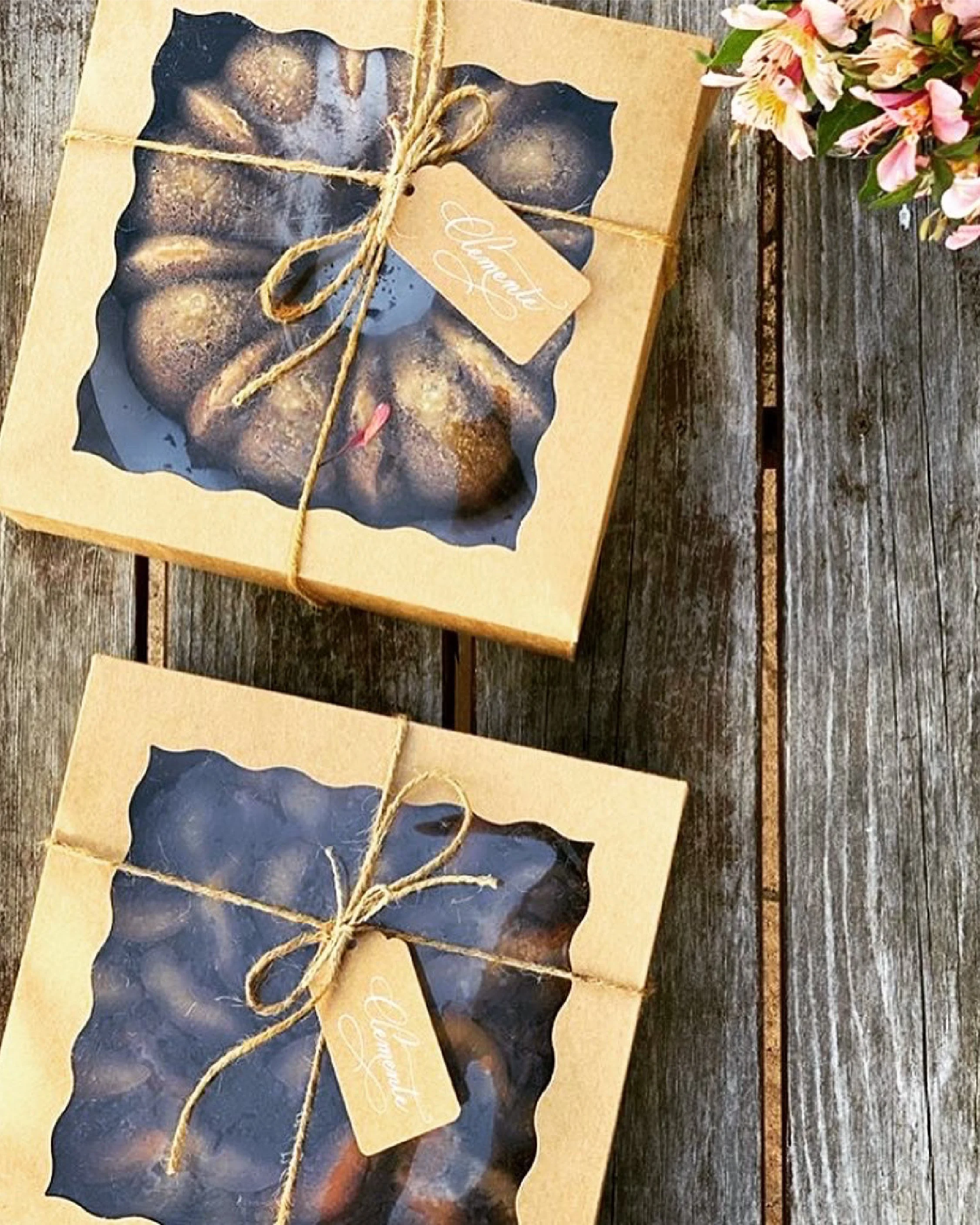 A gift box with a photo of blueberries on it, wrapped with twine and a beige tag that says 'Enjoy'. The box is placed on a rustic wooden table and there are pink flowers nearby.