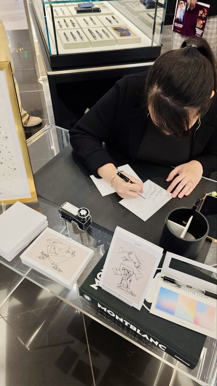 A woman is signing an autograph at a jewelry store display table with jewelry and stationery items, including Montblanc branded materials.