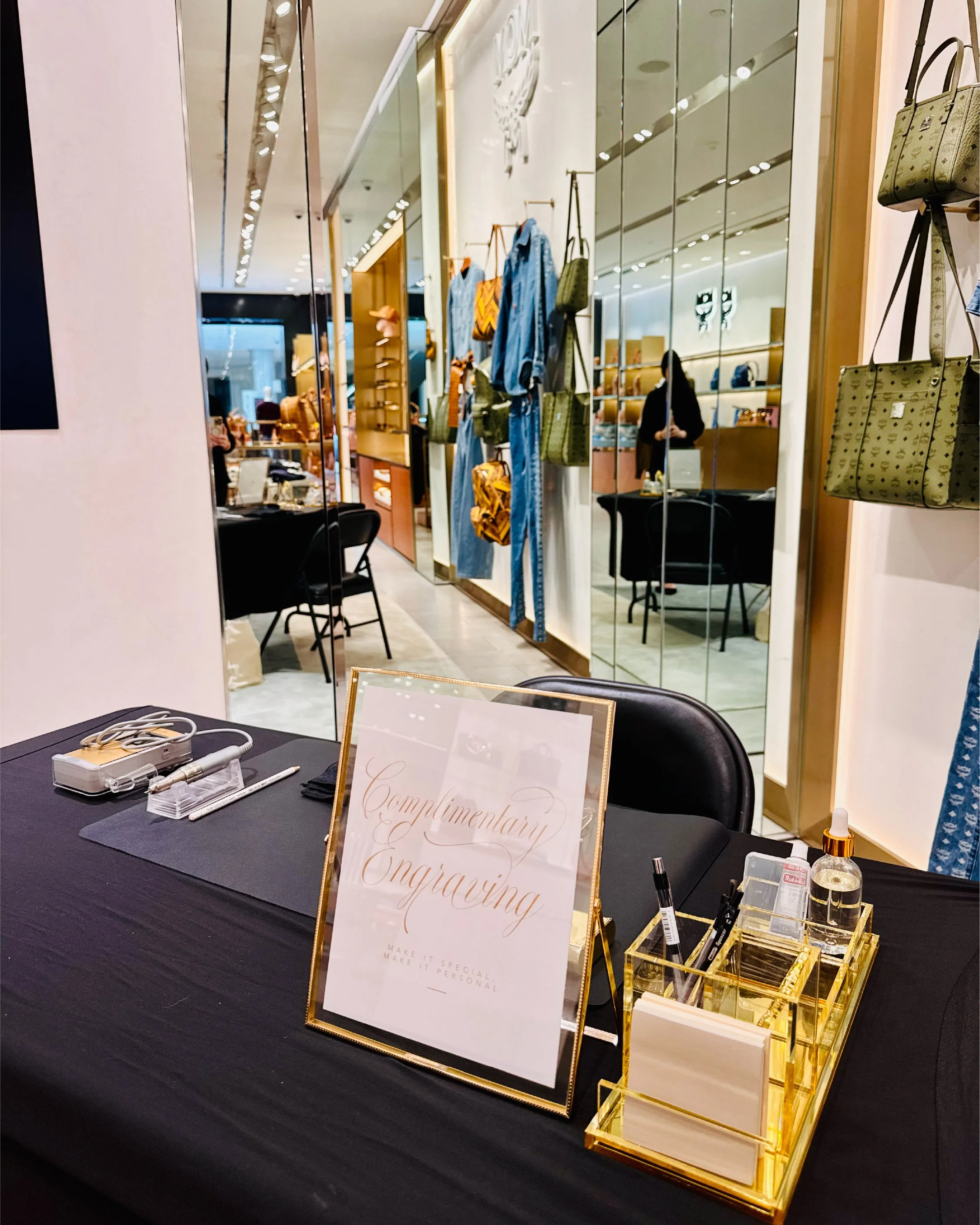 A retail display table with various makeup and skincare products, alongside a gold frame sign that says "Complimentary Engraving" and "Make it special, make it personal." The background features a clothing boutique with handbags hanging on the wall and a customer shopping.