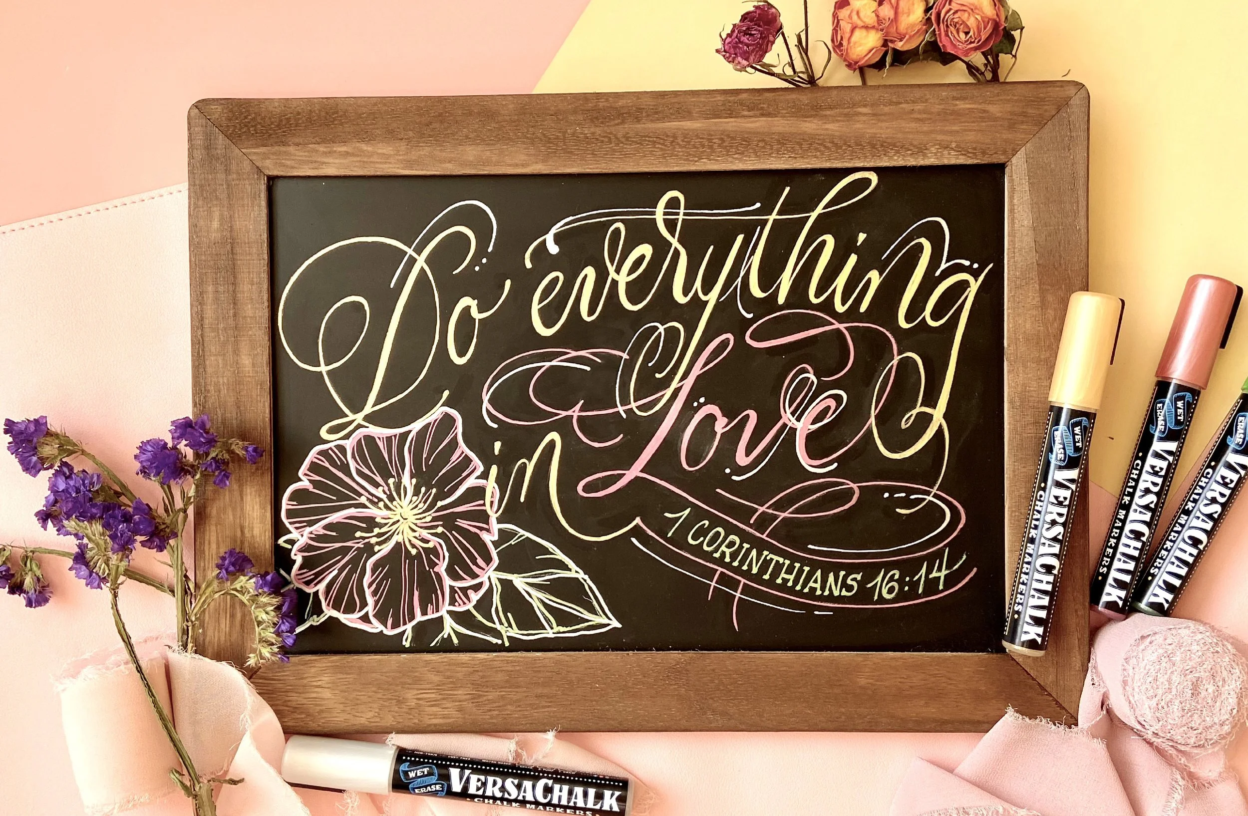 A decorative chalkboard sign with colorful lettering that reads 'Do everything in love' from 1 Corinthians 16:14, surrounded by dried purple flowers, pink fabric, and Four Different Colors of chalk markers, with a pink and yellow wall in the background.