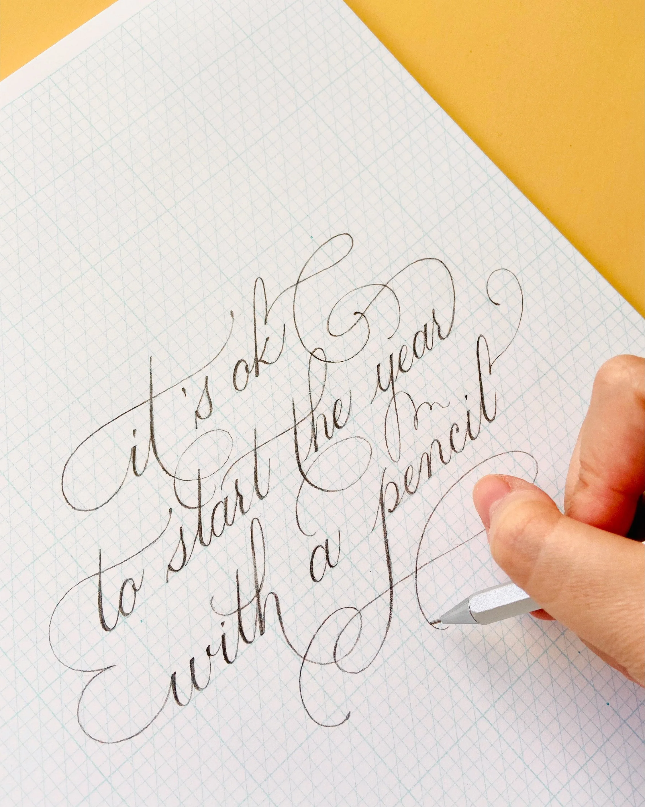 Handwriting in cursive on graph paper that says 'It's okay to wear what makes you happy' with a person's hand holding a pen in the bottom right corner.