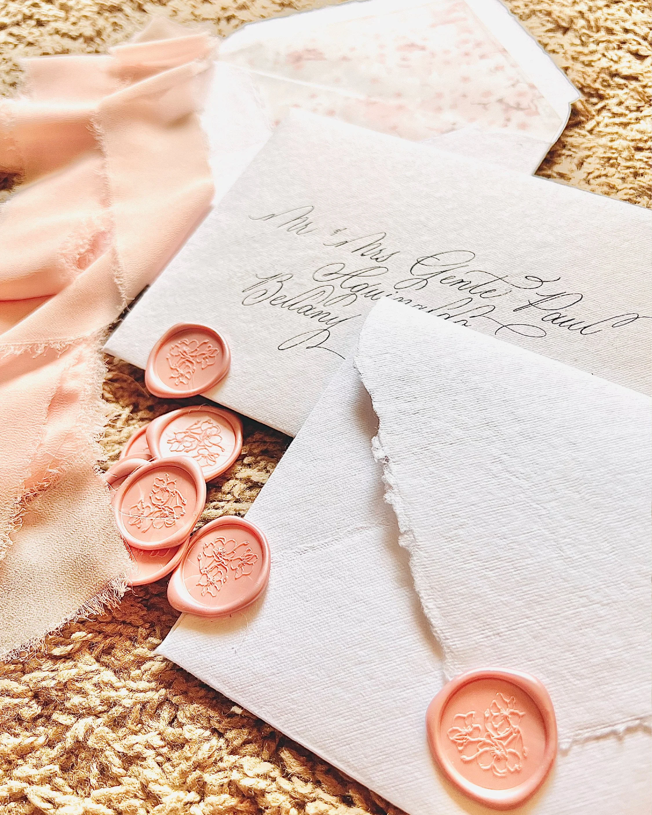 An envelope with handwritten calligraphy addressed to Mr. and Mrs. Gerard Laug, with a broken wax seal and pink wax seals with floral designs, placed on torn white paper on a woven surface.