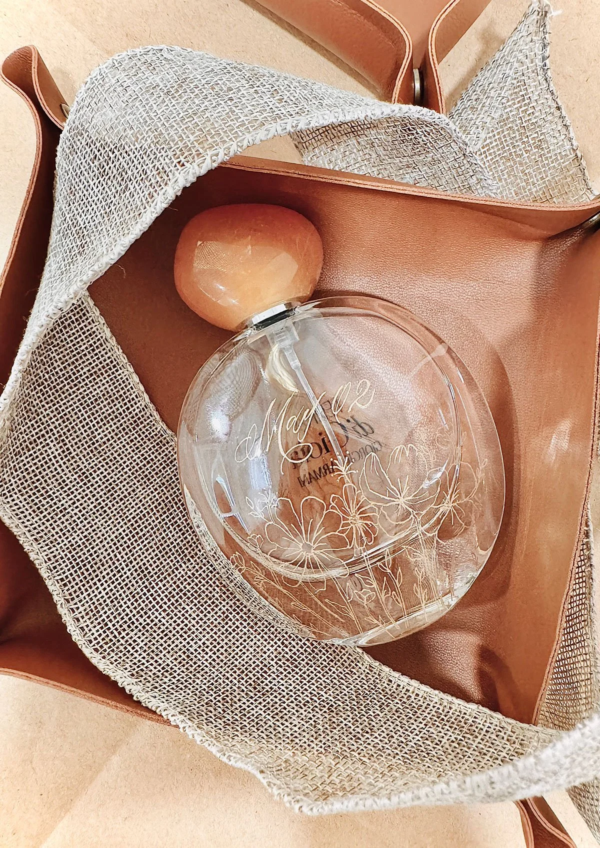 A clear glass perfume bottle with engraved floral designs and the words "May 02" on it, resting inside a leather and mesh-lined gift box.
