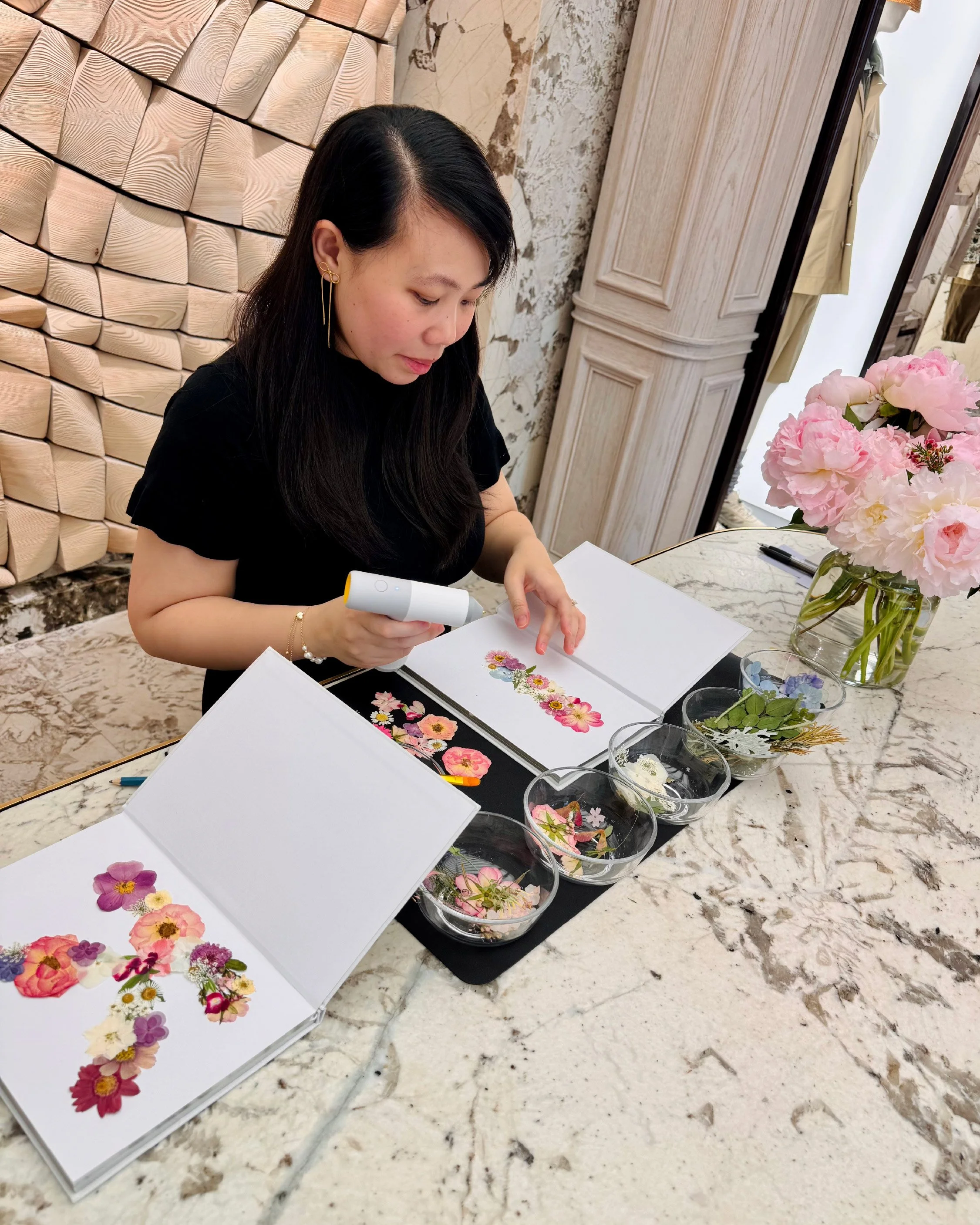A woman with black hair and gold jewelry is sitting at a table with flower illustrations, surrounded by various flowers and leaves, in a room with elegant decor, including a wooden panel wall and a flower arrangement on the table.