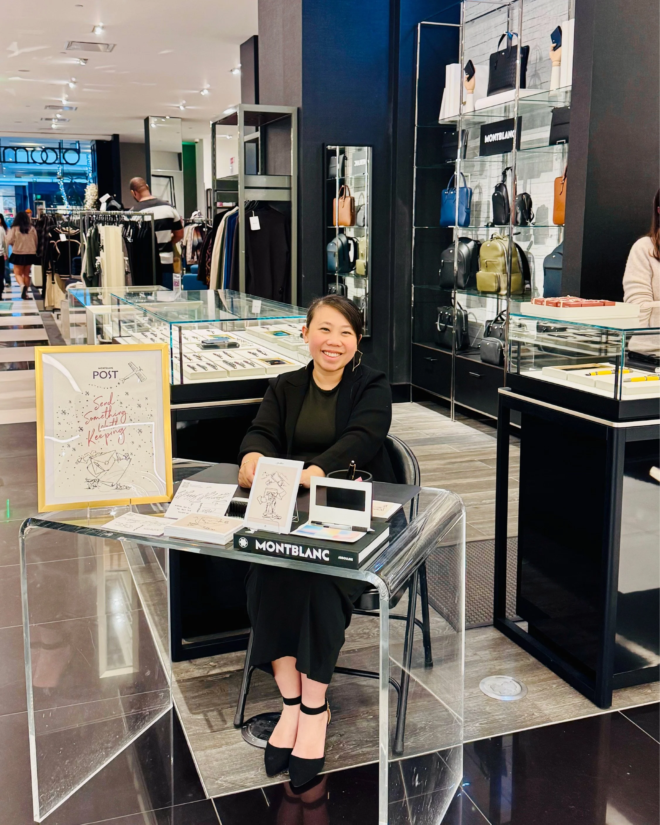 Smiling woman in black dress and black heels sitting at jewelry display table in store with handbags on shelves behind her.