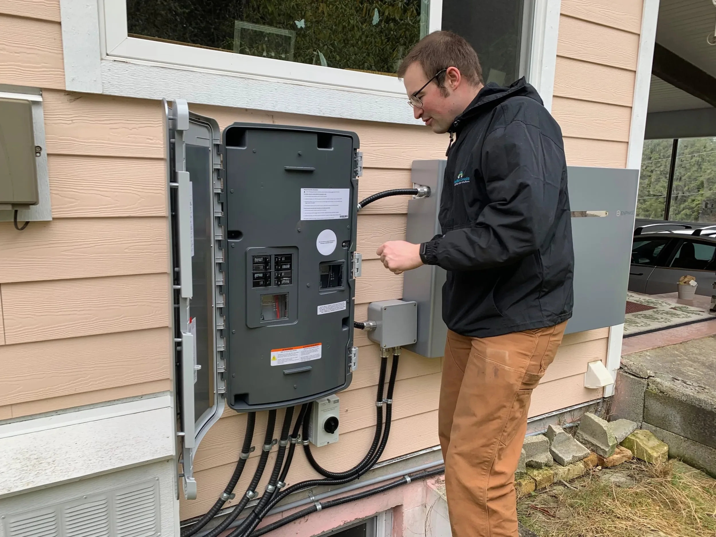 A man wearing glasses and a black jacket working on an electrical panel outside a house with beige siding and white trim.
