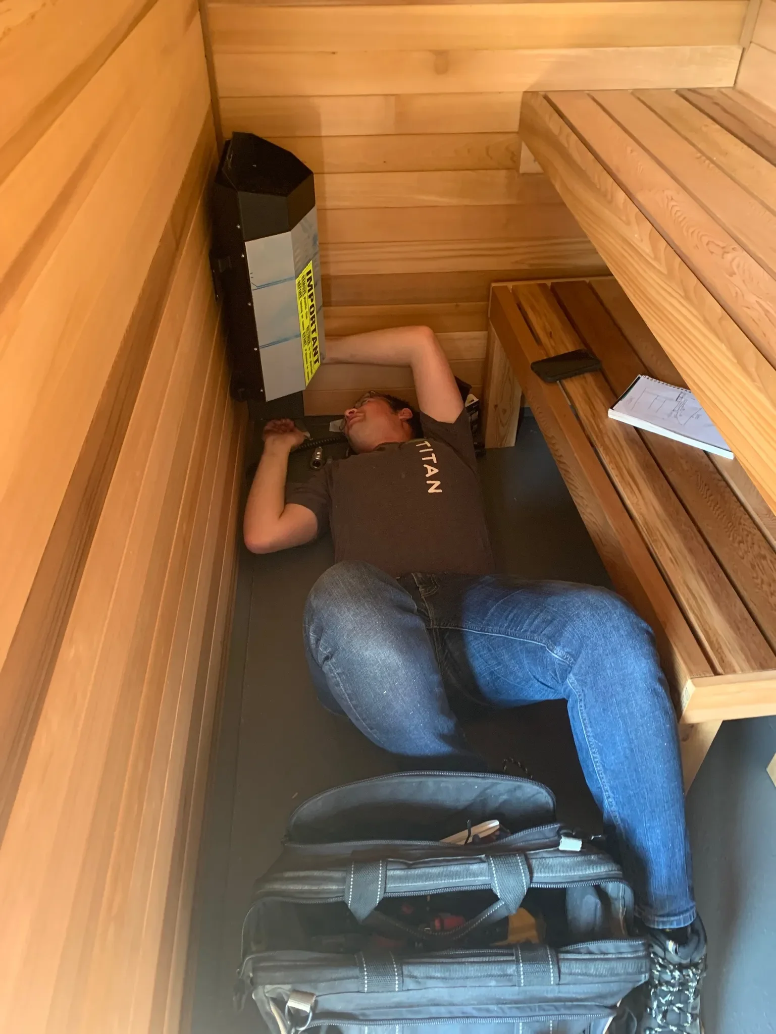 A man with glasses and a beard, lying down on his back in a small wooden room, is talking on a landline phone. There are wooden benches and a backpack in the room. The man is wearing a dark shirt and jeans.