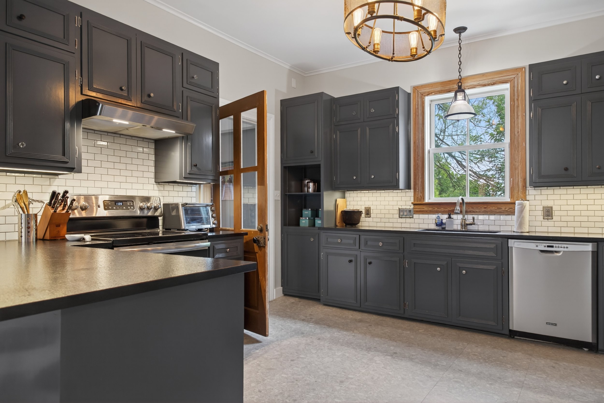 Kitchen with dark gray cabinets, white brick backsplash, stainless steel stove, large window with wooden trim, and a ceiling light fixture.