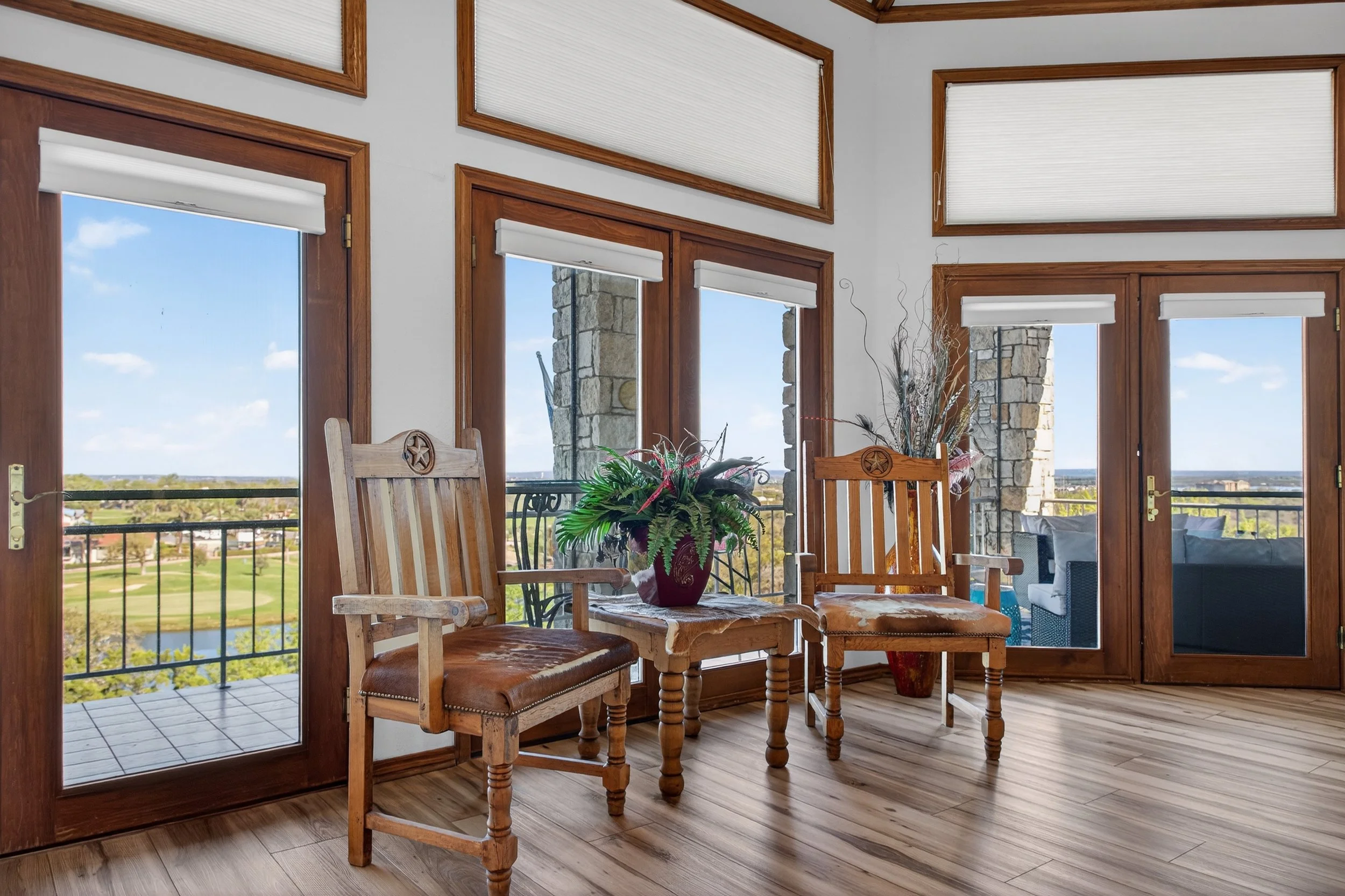 A dining area with wooden chairs and a table, a large potted plant, and a view of a golf course through glass doors and windows with white blinds, brick accents, and a balcony outside.