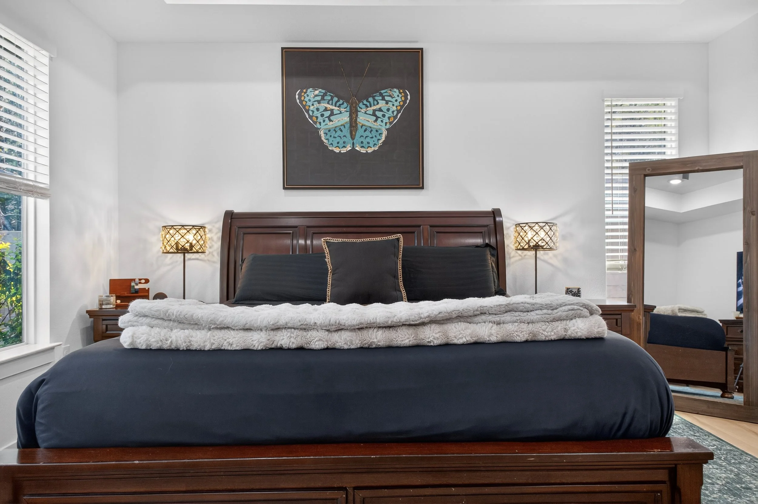 A bedroom with a large wooden bed, black bedding, and a white furry blanket. Two matching lamps on bedside tables, a framed butterfly artwork on the wall, and windows with white blinds.