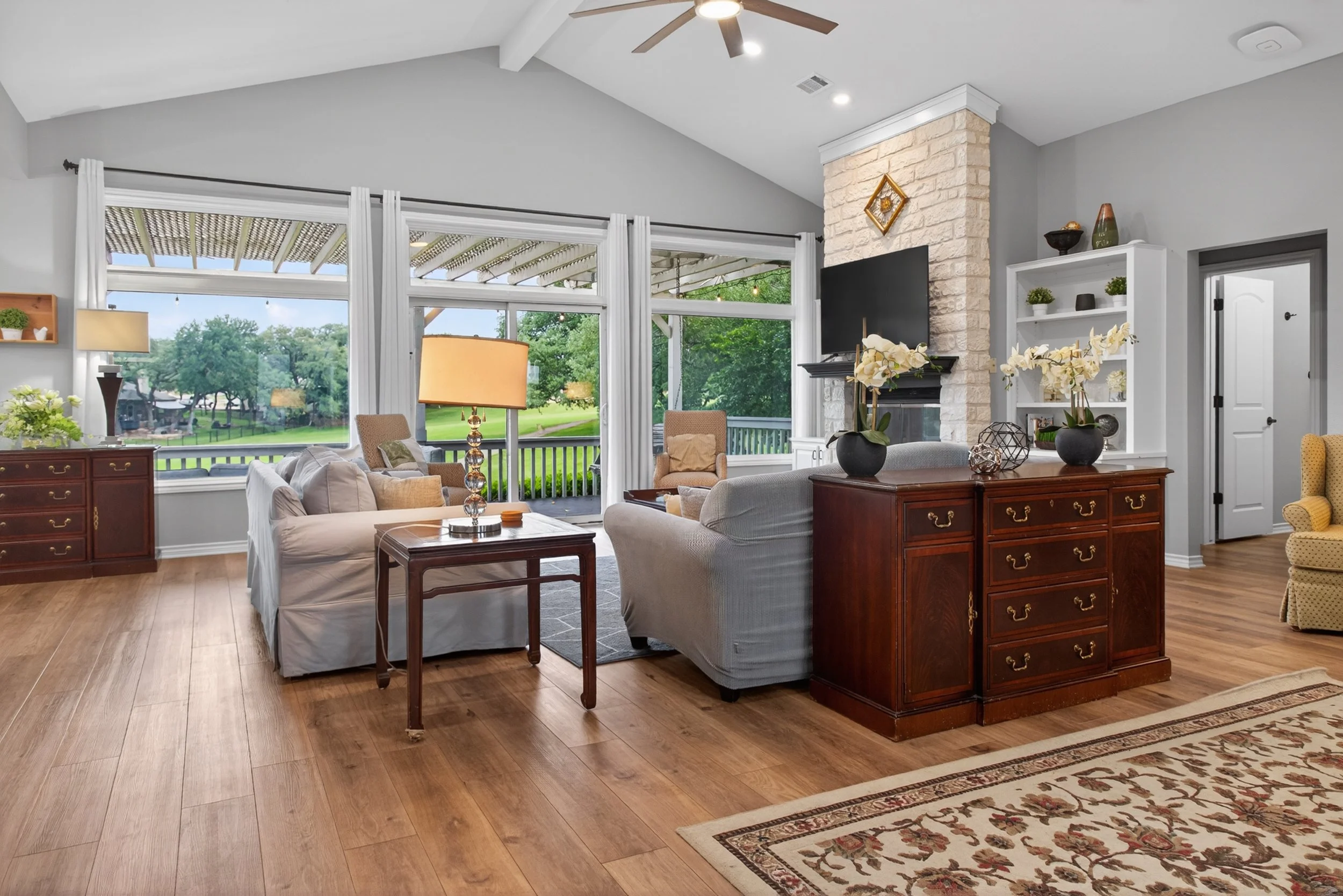 Living room with large windows, ceiling fan, and fireplace. Furniture includes sofas, armchairs, and wooden tables. Decor features lamps, vases, and plants. Outside view shows trees and a porch.