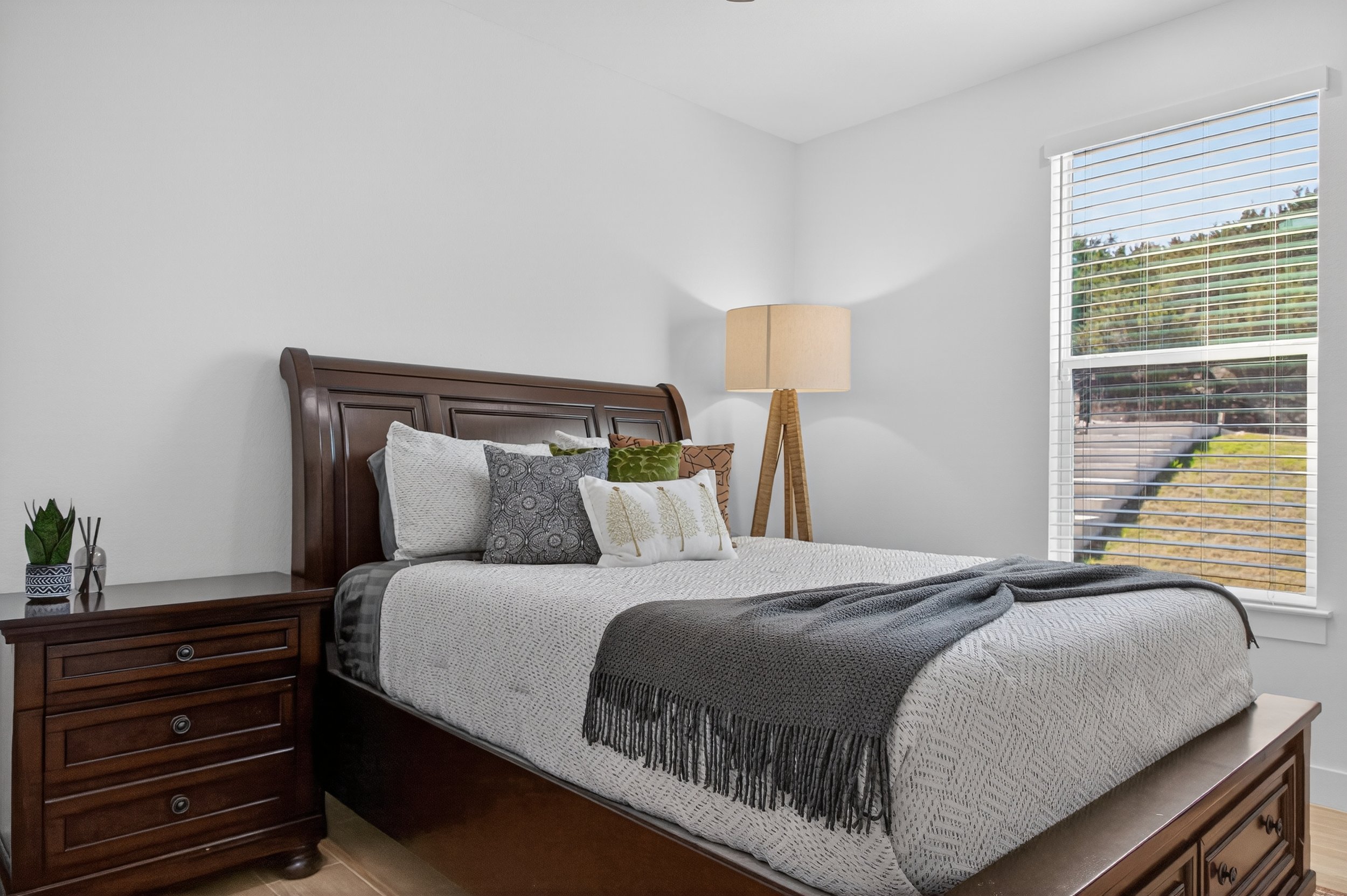A neatly made bed with pillows, a dark gray blanket, and a wooden headboard in a room with white walls and a window showing a green outdoor scene. There is a nightstand with a star-shaped plant and a lamp next to the bed.