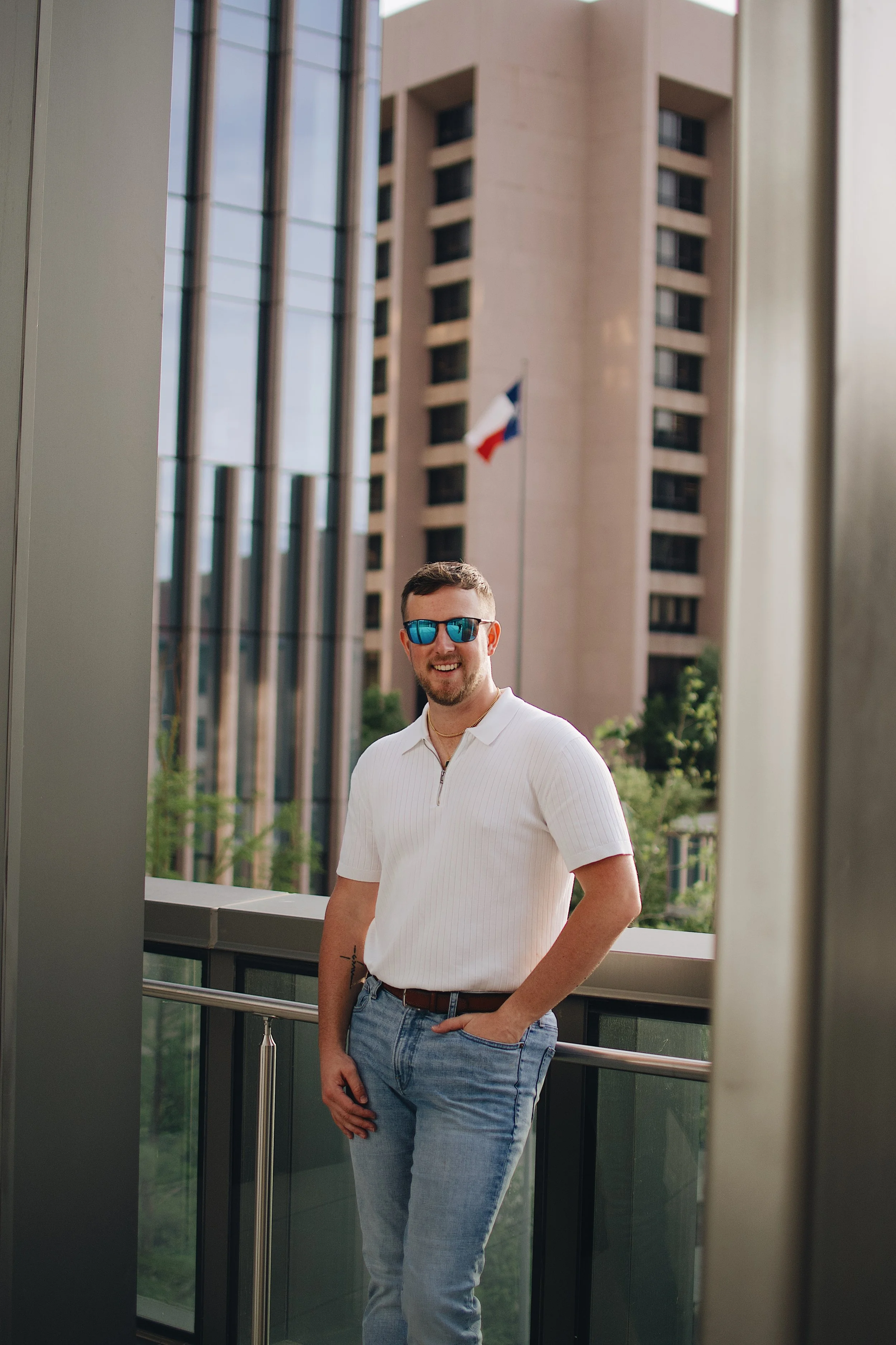 A man with short hair, a beard, and sunglasses is standing on a balcony, smiling. He is wearing a white half-zip shirt and light blue jeans. Behind him, there is a building with multiple floors, and a flag is visible in the background.
