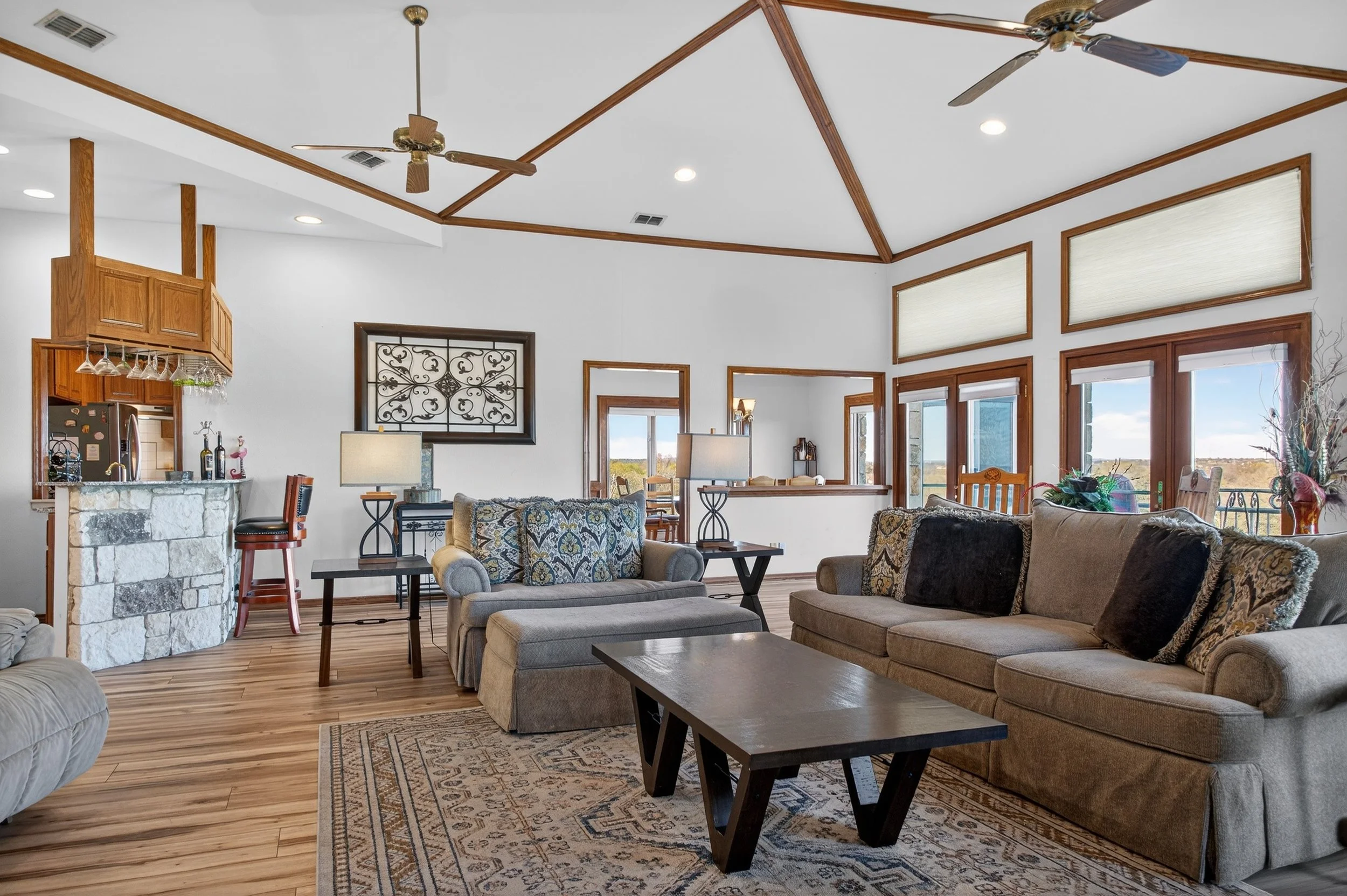 Living room with a beige sectional sofa, patterned armchair, wooden coffee table, and large windows showing an outdoor view, with ceiling fans and wood trim accents.
