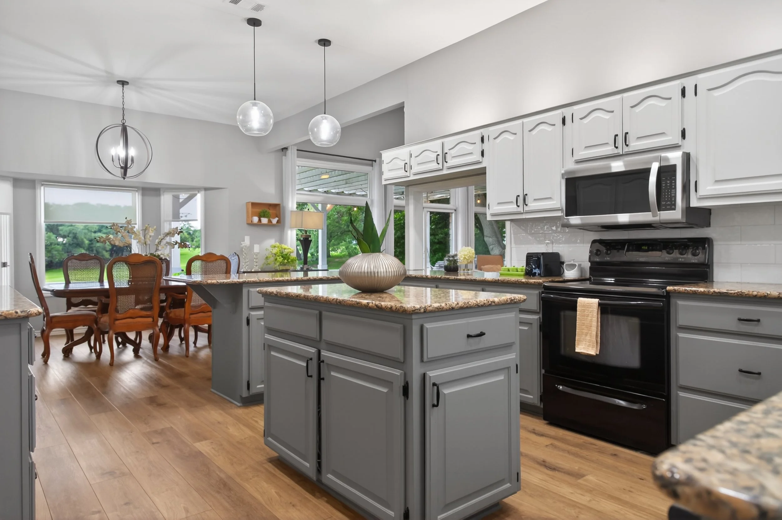 Modern white kitchen with granite countertops, island, black stove, microwave, and white cabinets, adjacent to a dining area with wooden chairs and a large window showing greenery outside.