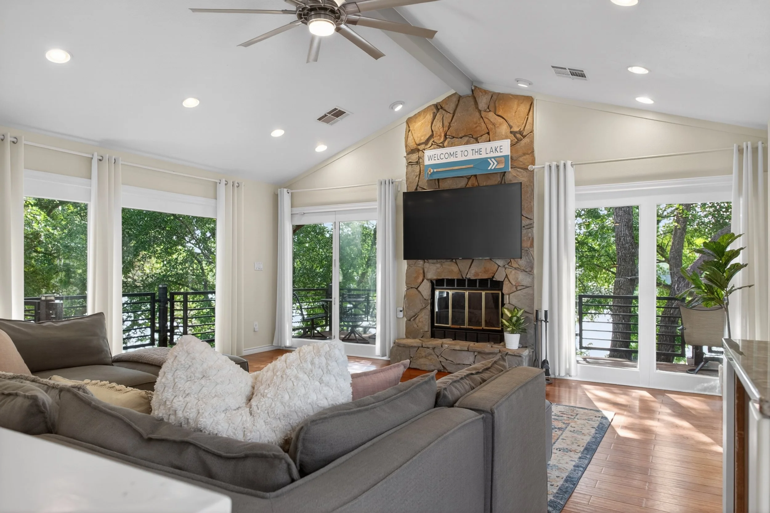 Living room with a gray sectional sofa, a stone fireplace with a mounted TV, large windows showing lush green trees, white curtains, and a ceiling fan.
