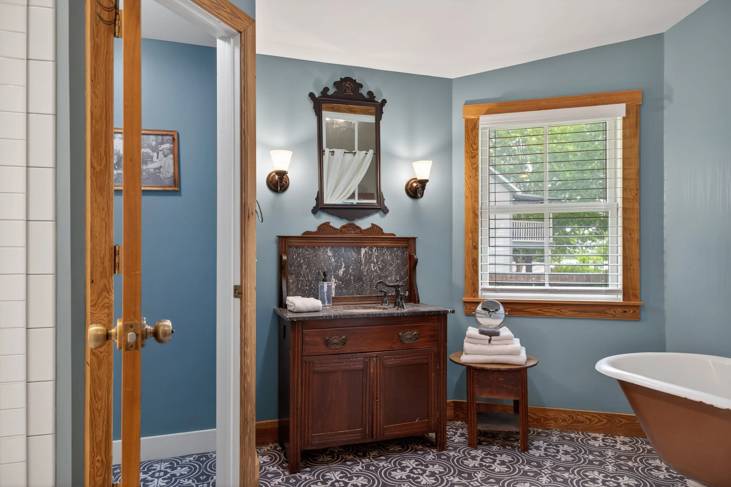 A bathroom with blue walls, a window with wooden trim, a wooden vanity with a marble top, and a mirror flanked by two wall sconces. The bathroom has patterned black and white floor tiles.
