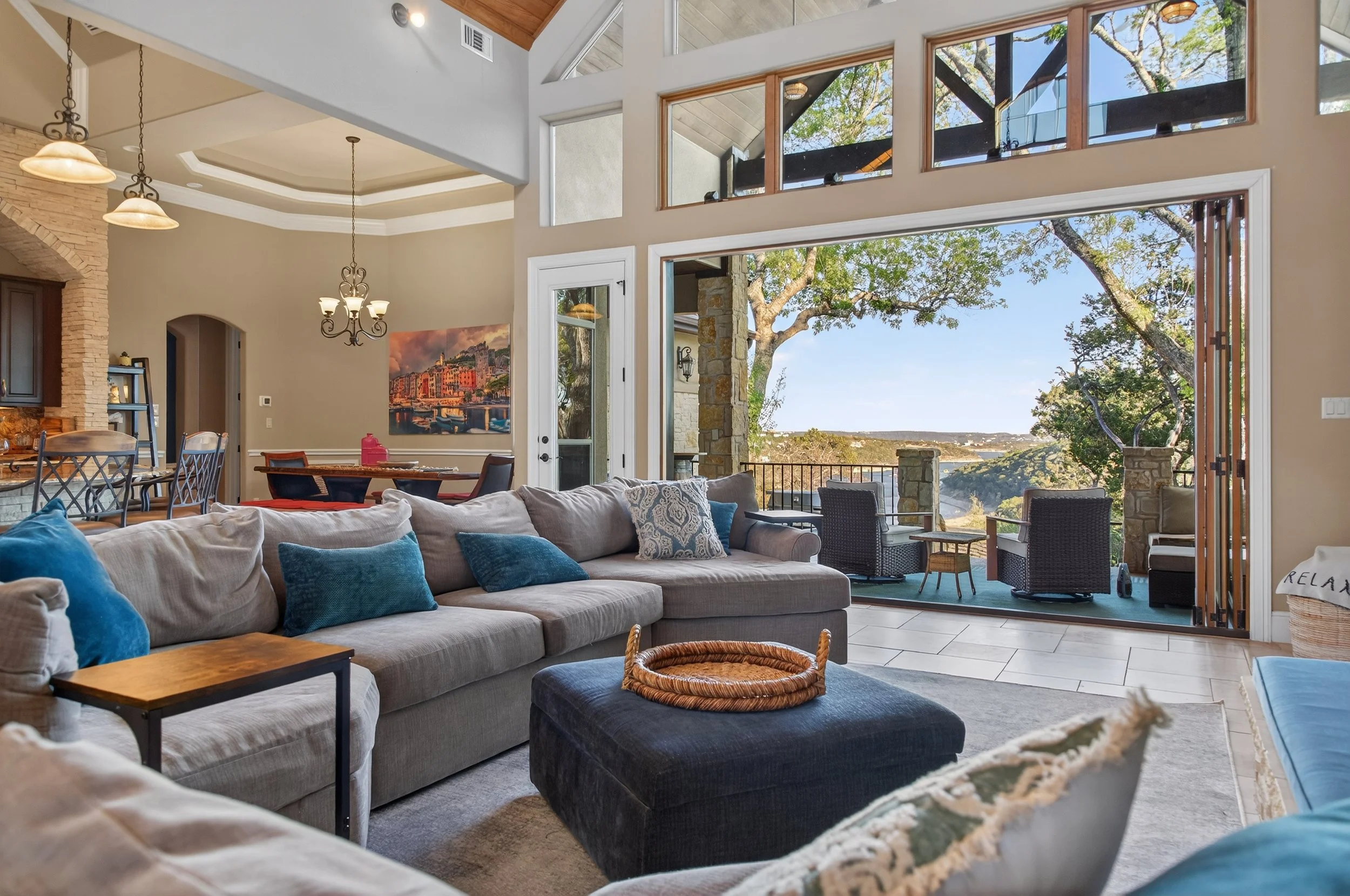 Spacious living room with large windows overlooking trees and a scenic outdoor view, beige walls, a light-colored sectional sofa with blue and patterned pillows, a black ottoman with a woven tray, a dining area with a wooden table and chairs, a stone fireplace, and ceiling fixtures.