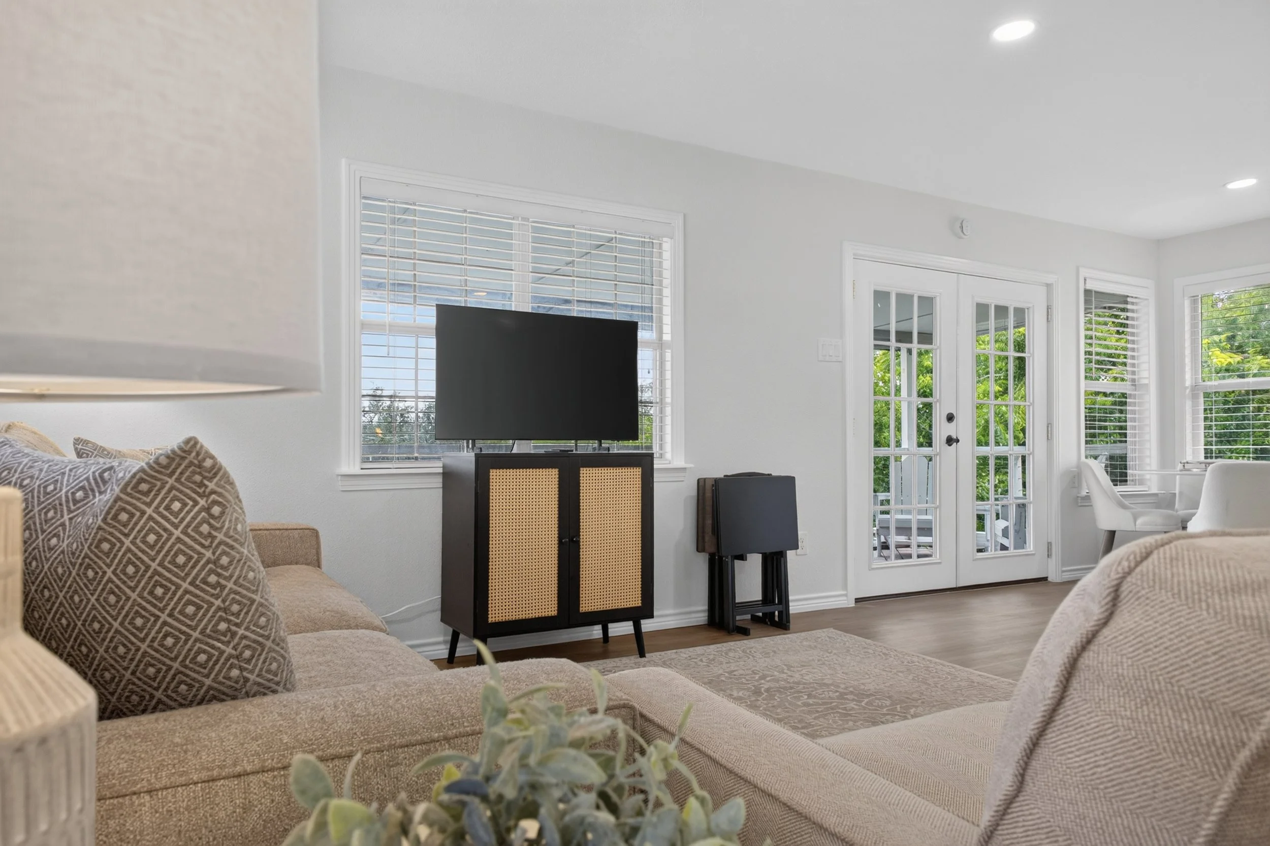 Living room with beige sectional sofa, patterned pillows, a flat-screen TV on a stand, large windows with blinds, and a glass door leading outside, with greenery visible through the windows.