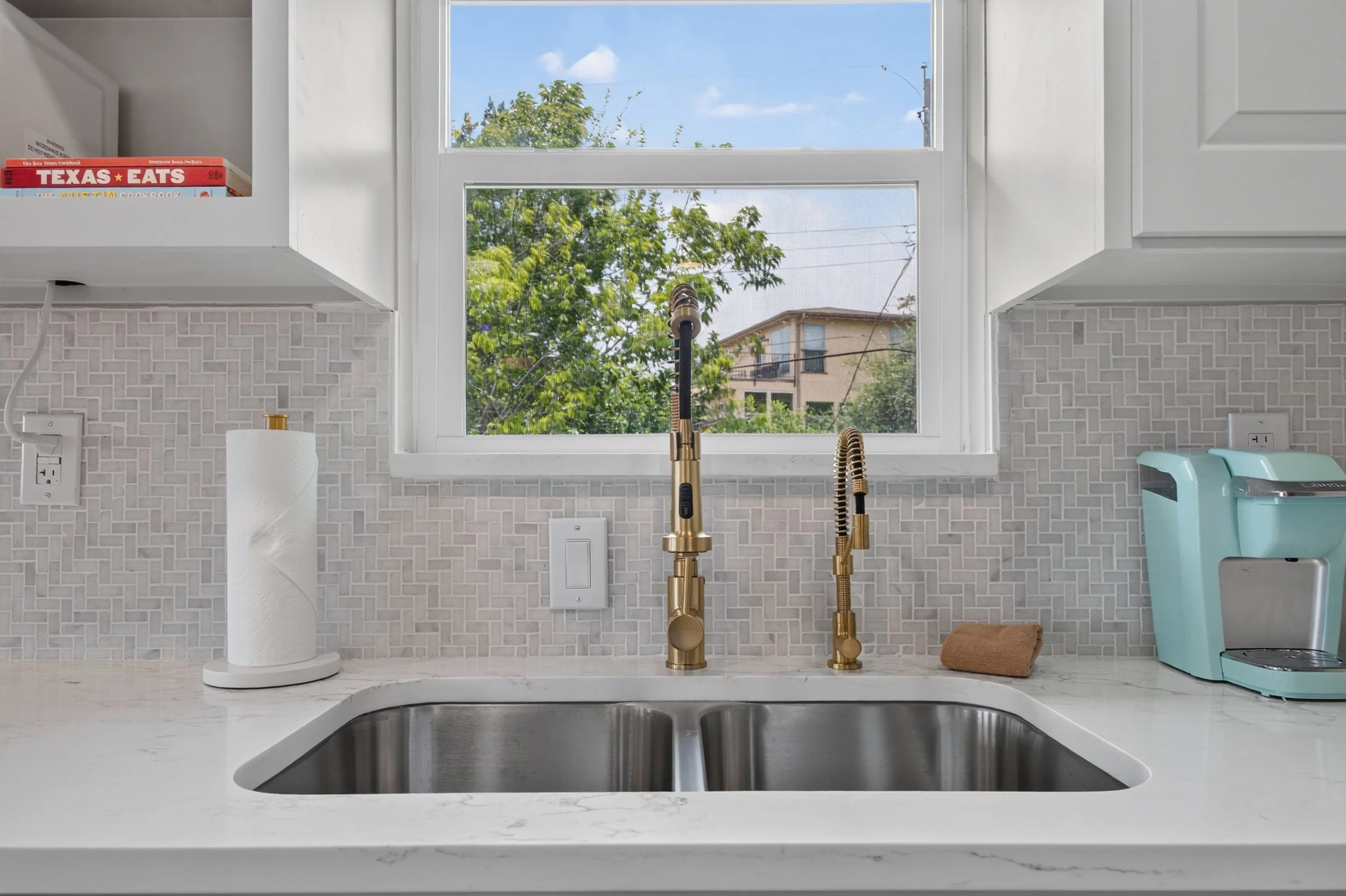 Kitchen sink area with a window showing trees and a building outside. The countertop has a paper towel roll, a faucet, and a small appliance, possibly a water filter. There are kitchen cabinets above and a backsplash with a geometric tile pattern.