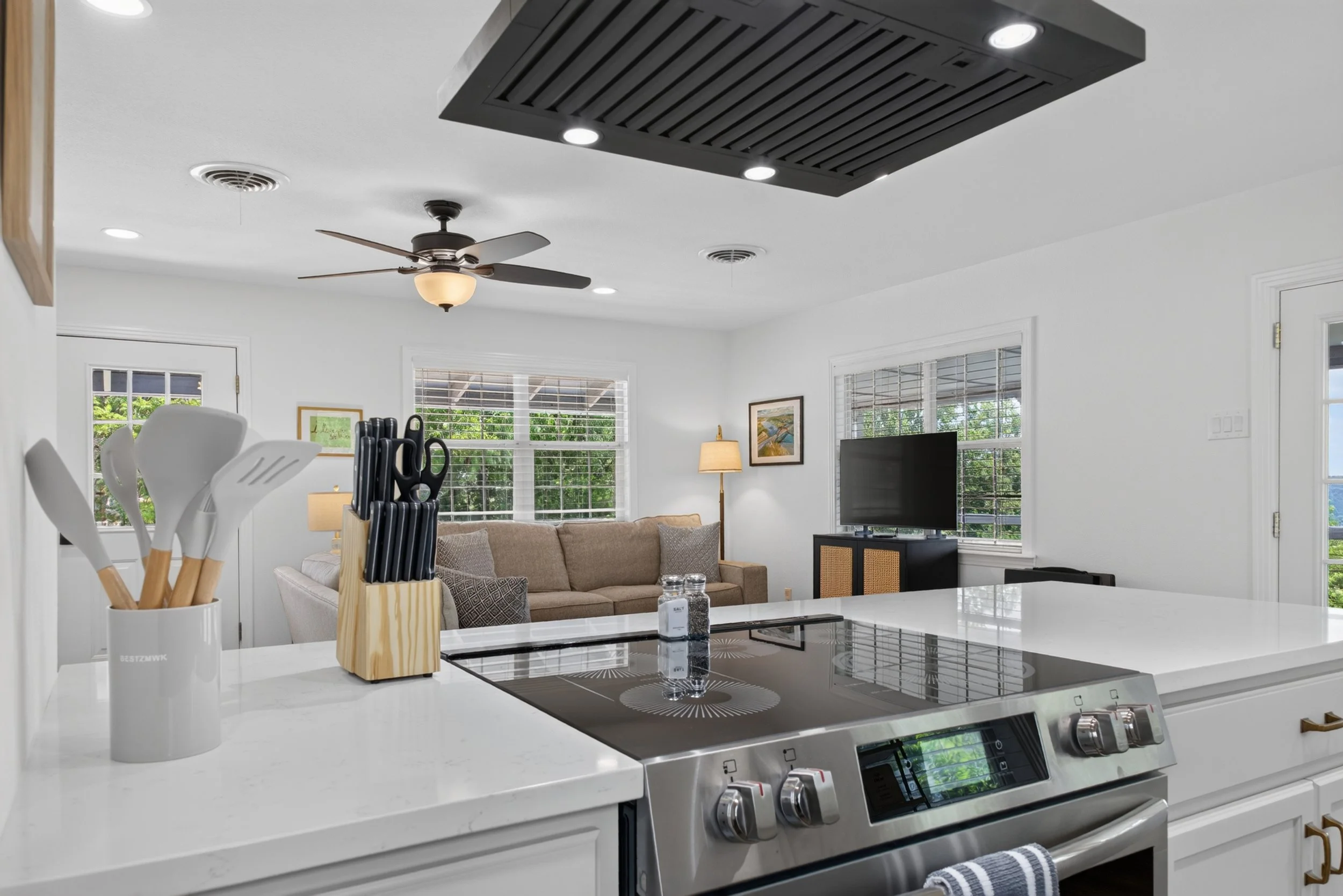 Modern kitchen with white countertops and cabinetry, black stove, utensil holder, and a view of a living room with brown sofa, window, TV, and ceiling fan.