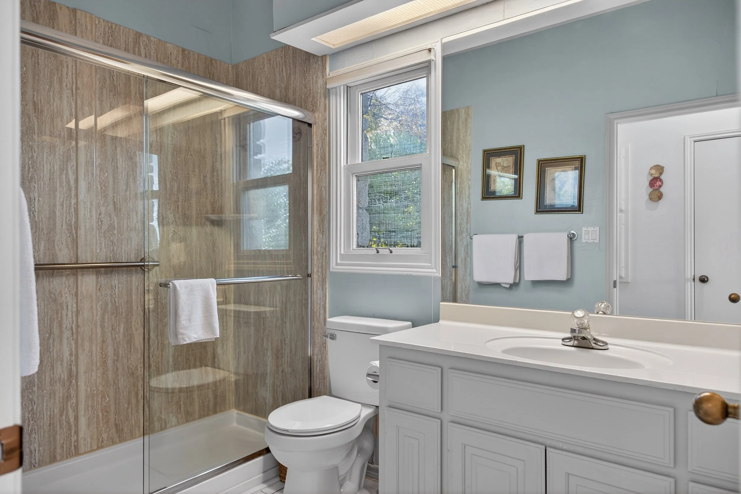 Bathroom with light blue walls, a window with blinds, a white sink vanity with cabinet storage, a toilet, and a walk-in shower with beige tiles and a glass door.