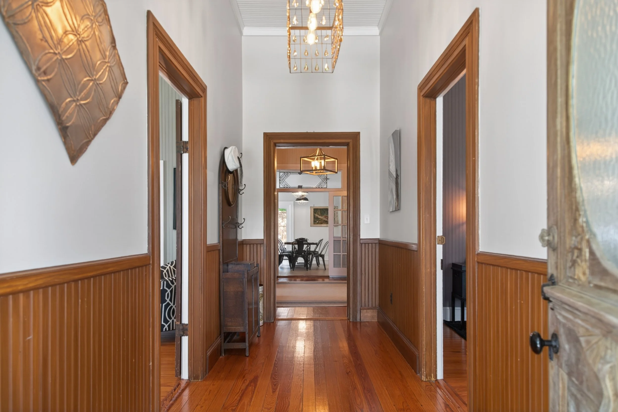 A hallway with wood accents and three doorways leading into rooms. There is a chandelier hanging from the ceiling and artwork on the walls.