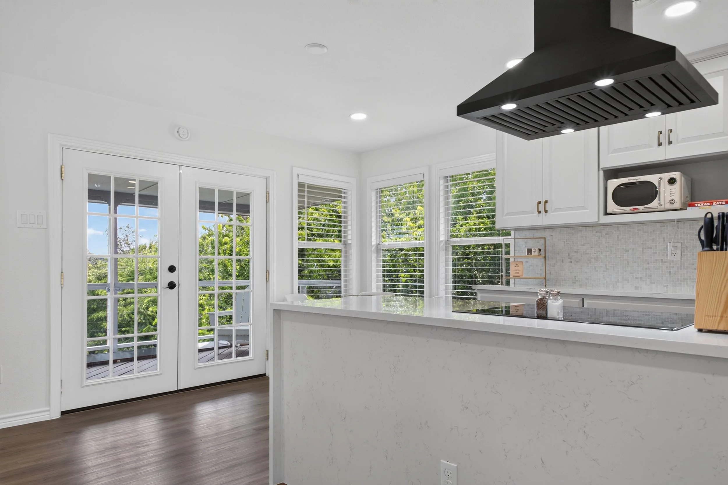 A bright kitchen with white cabinets, a marble island, large windows with white blinds, and a door leading outside. There is a black range hood with built-in lights above the kitchen island.