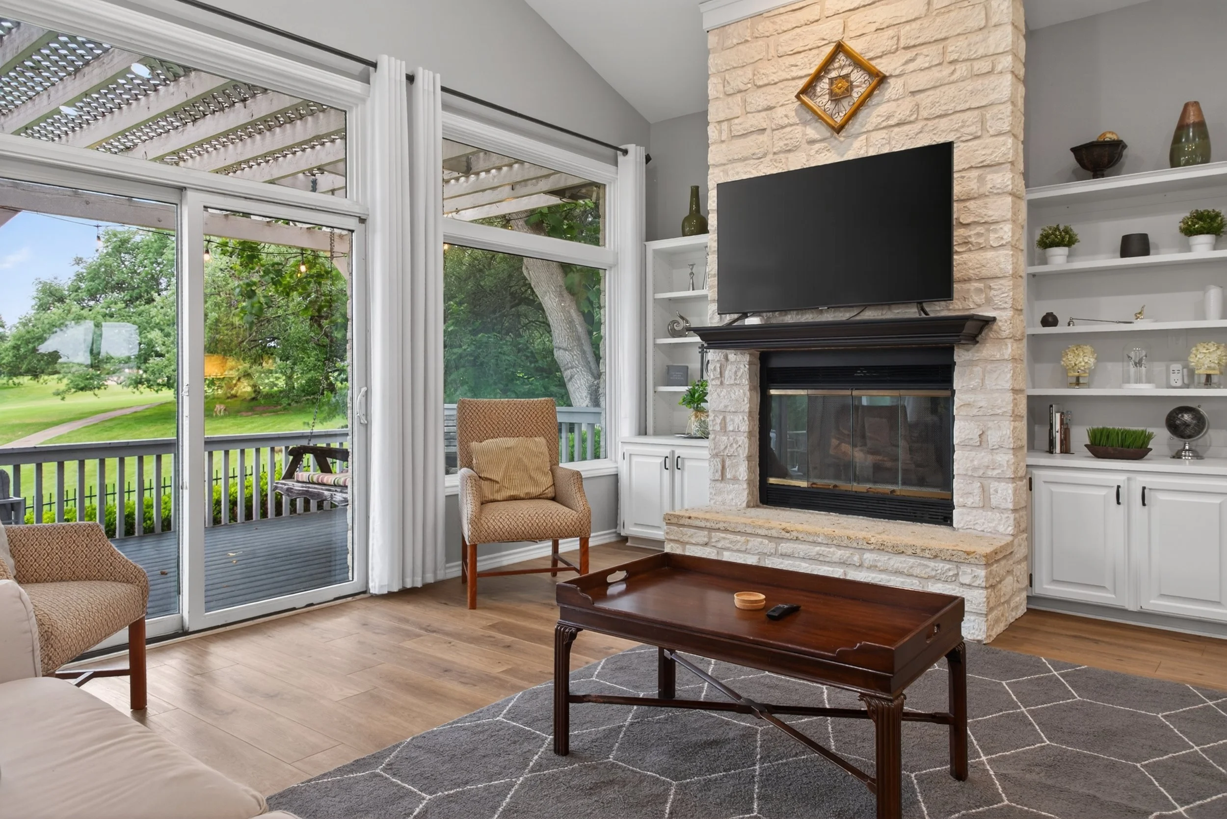 Living room with large sliding glass door showing a backyard with trees and grass, beige armchair near the door, white shelves on the wall with decorative items, a stone fireplace with a mounted TV above, and a wooden coffee table on a gray rug.