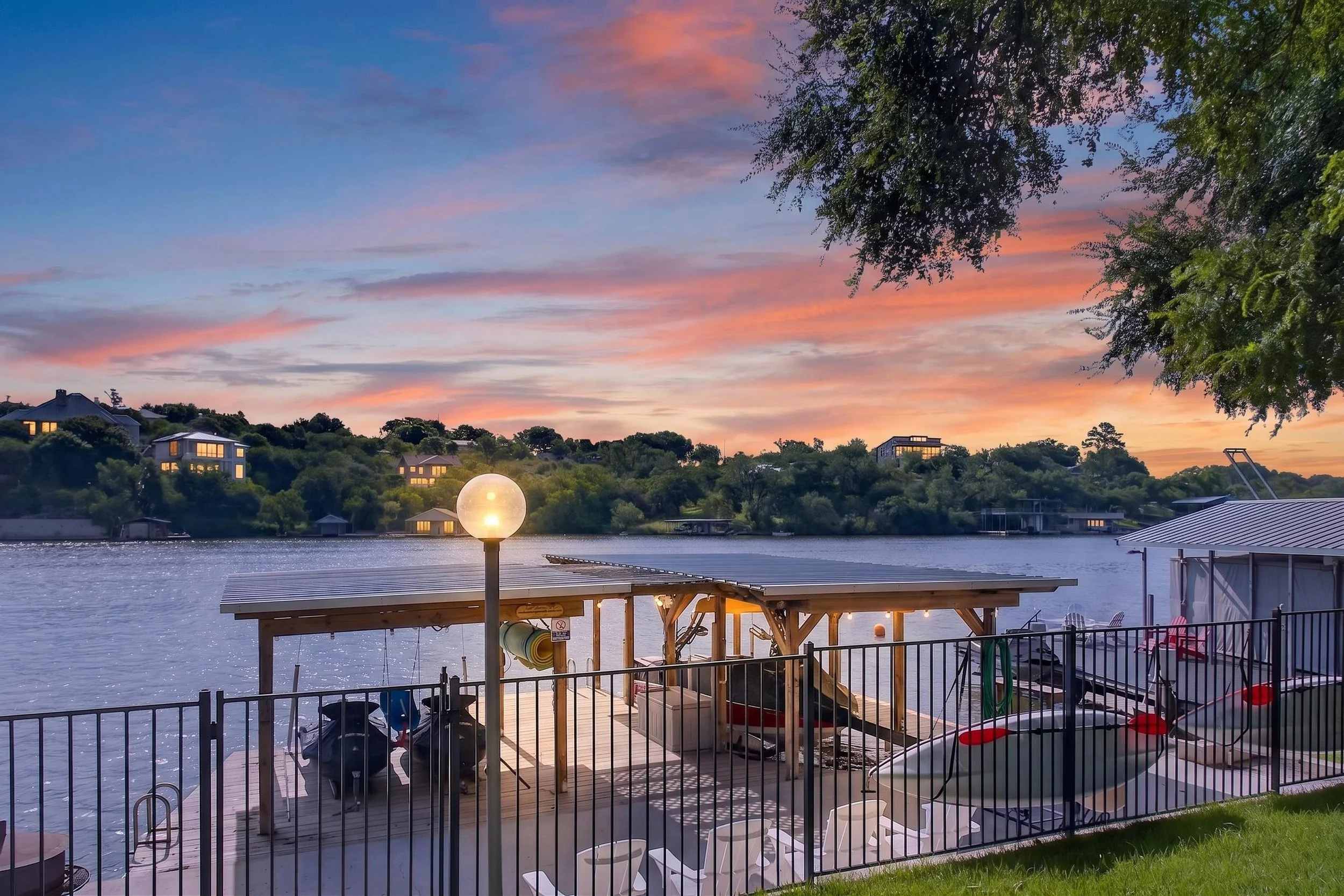 A lakeside scene at sunset with colorful pink and blue sky, houses on a hillside, and a waterfront area with a fenced deck, tables, chairs, and a lamppost near the water.