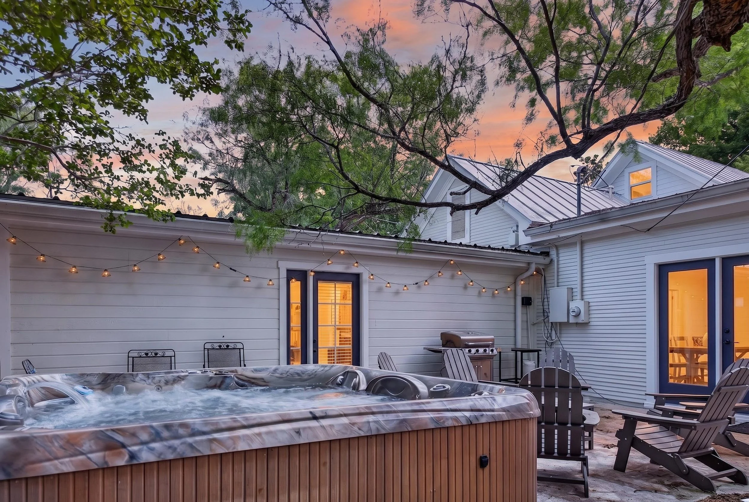 Backyard patio with hot tub, outdoor seating, string lights, and view of house with large windows, surrounded by trees during sunset.