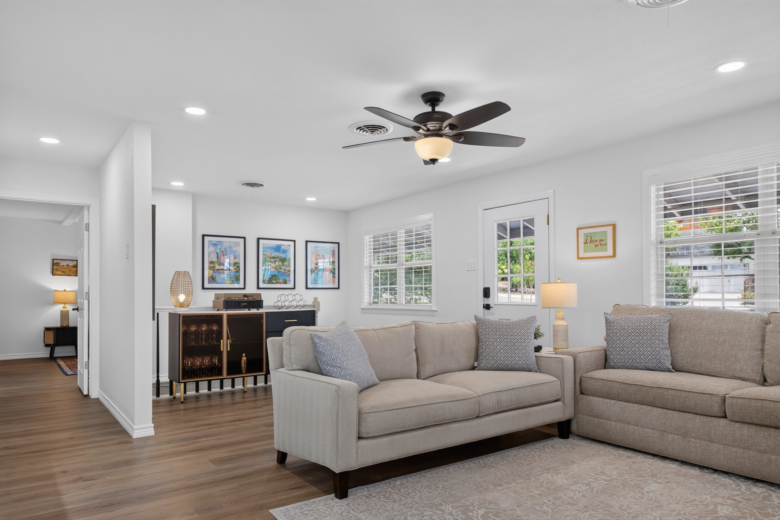Living room with beige sectional sofa, patterned pillows, large windows with white blinds, dark wooden floor, ceiling fan with light, artwork on the wall, and a small cabinet with decor items.