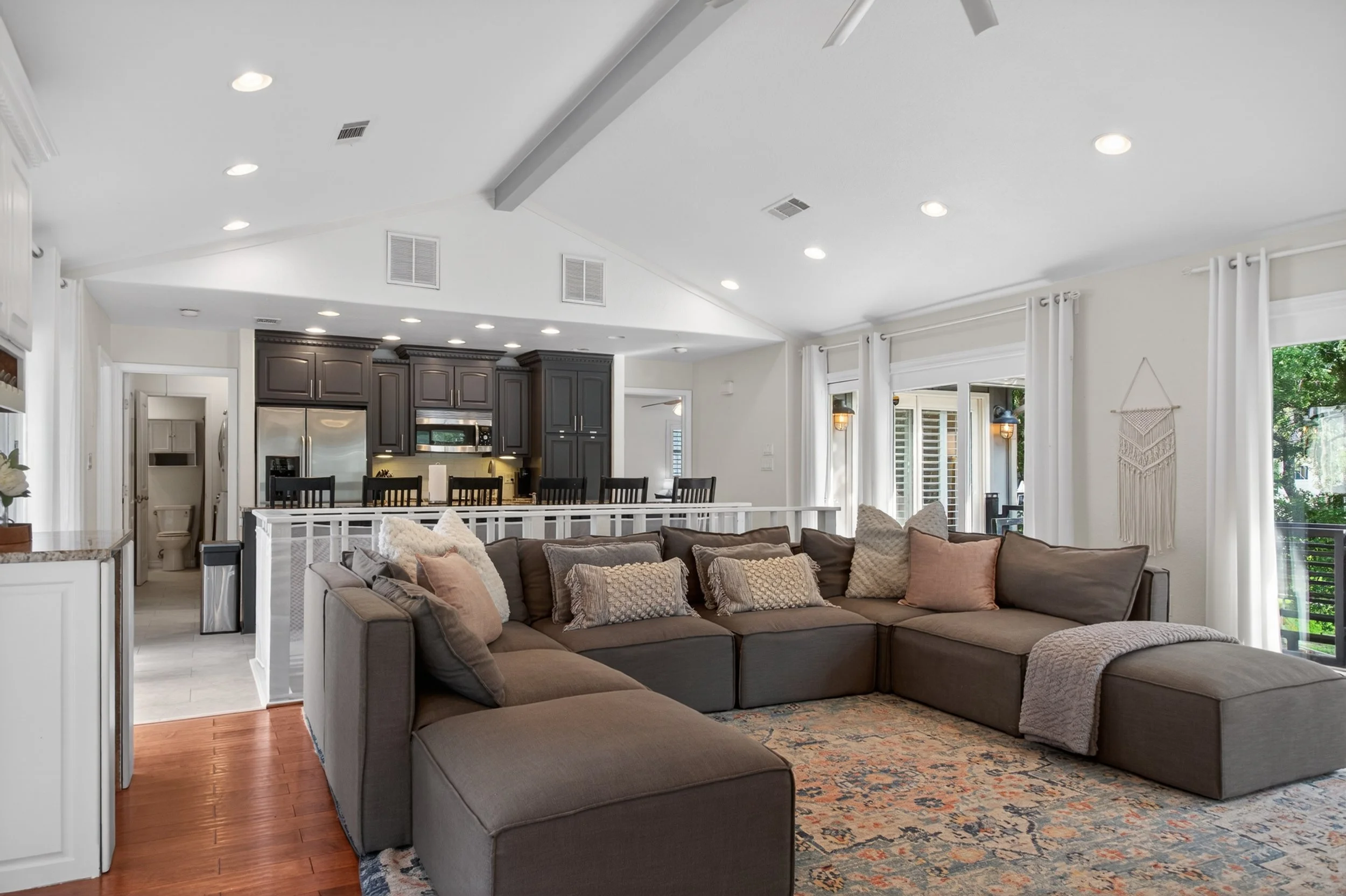 Spacious living room with large sectional sofa, multiple pillows, and a patterned area rug. Open kitchen and dining area with dark cabinetry, stainless steel appliances, and a breakfast bar. Bright room with large windows, white walls, and hanging wa