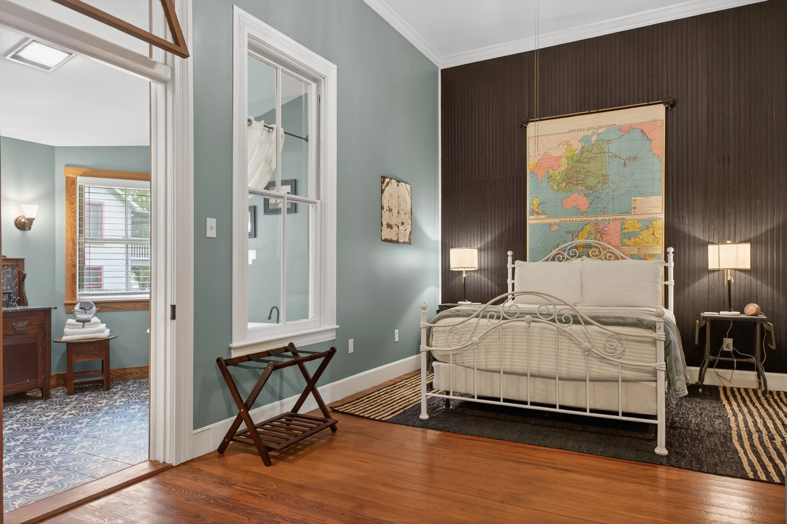 A bedroom with light blue walls, a white metal bed with a quilt, a dark wood accent wall with a large world map, two bedside lamps, a window with blinds, and a wooden floor.