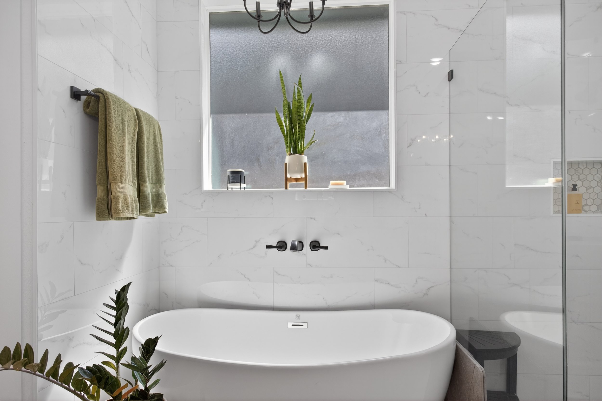 Modern bathroom with white marble walls, a bathtub, a frosted window with a potted plant, beige towels on a black wall hook, and various toiletries on the windowsill.