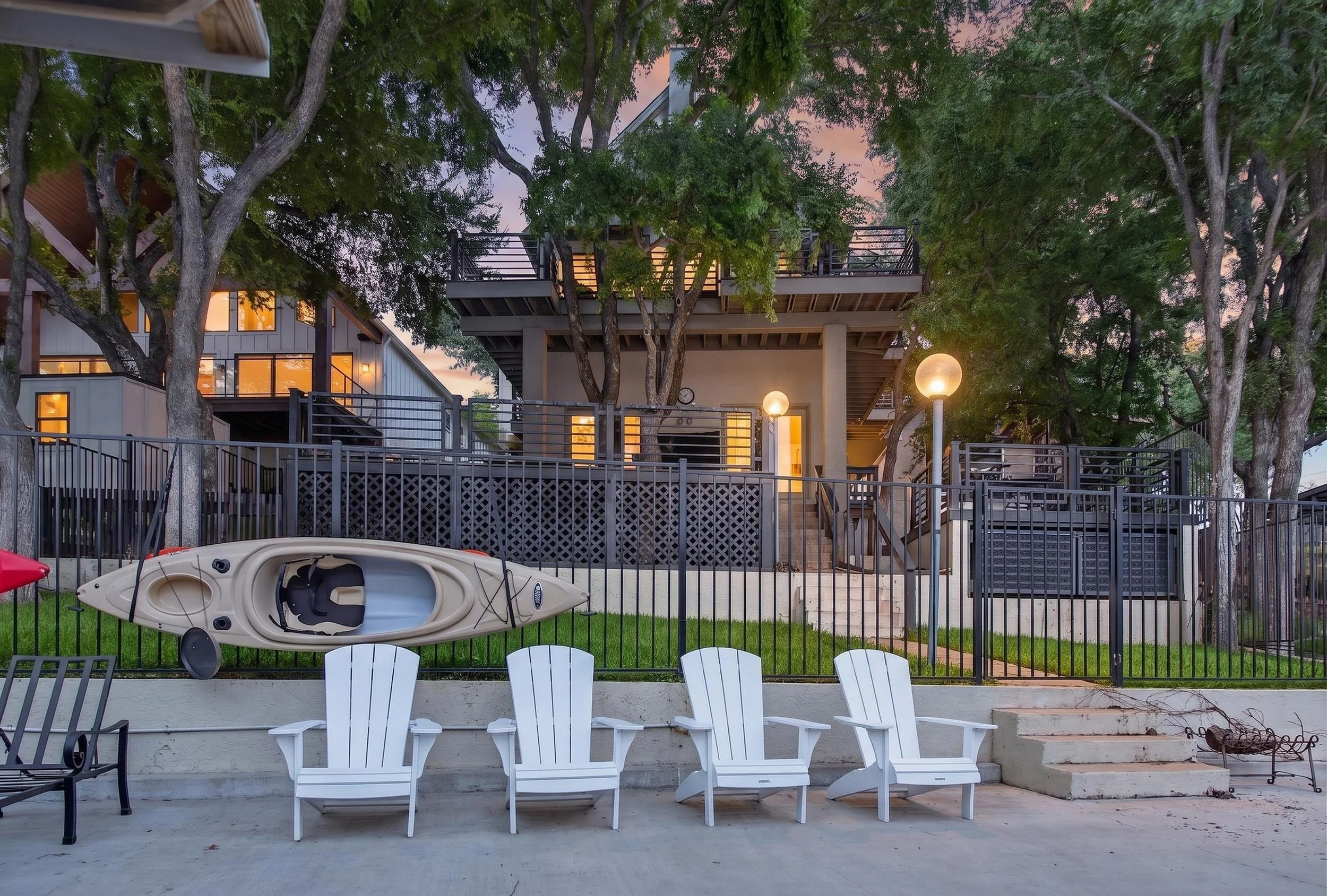 Backyard patio area with white chairs, a kayak, and a fenced yard, with a multi-story house with large windows and outdoor lighting surrounded by trees at dusk.