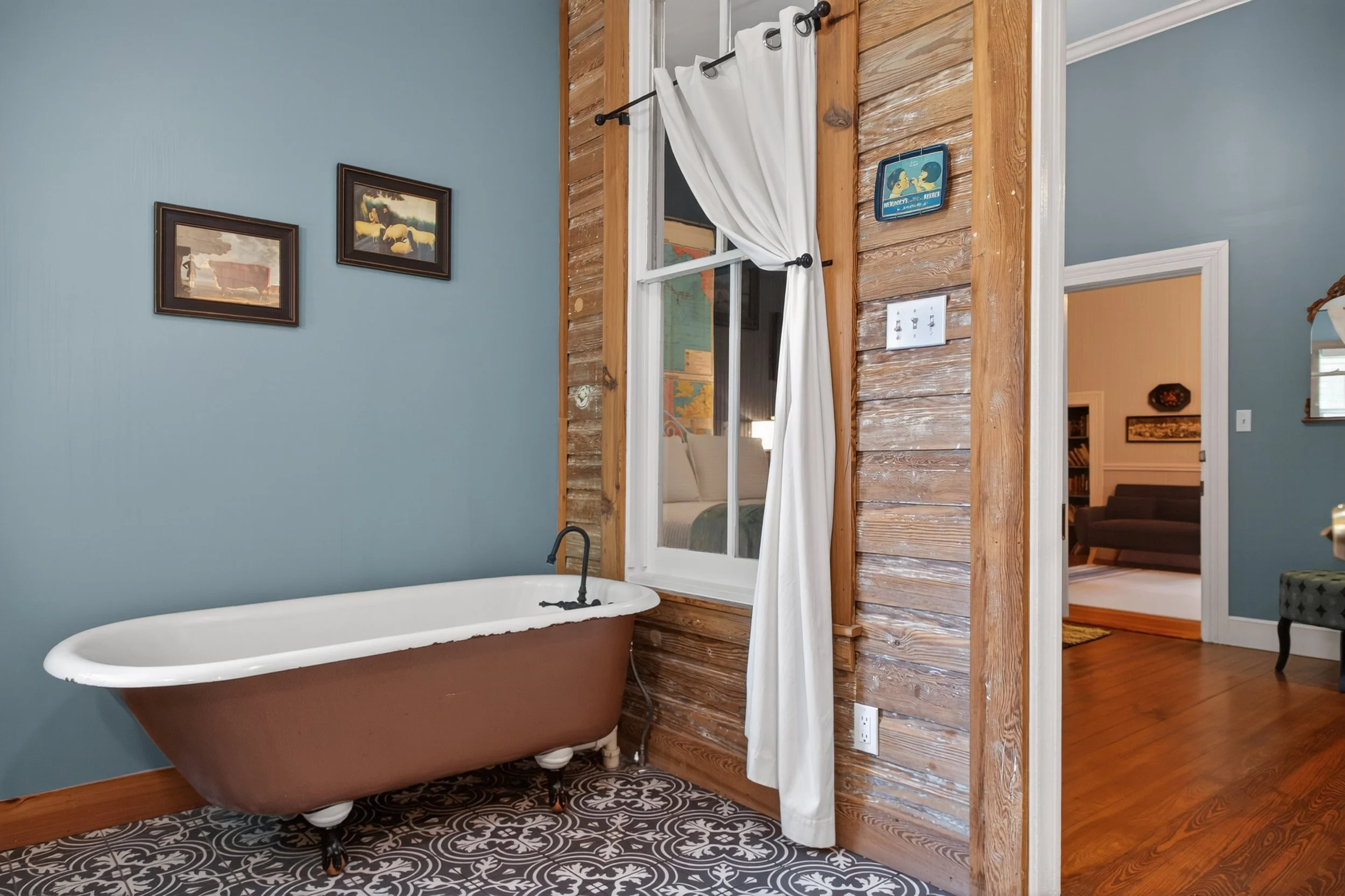 A rustic bathroom with a freestanding clawfoot bathtub against a blue wall, and a wooden partition with a window and a white curtain, connecting to a living room with a brown sofa.