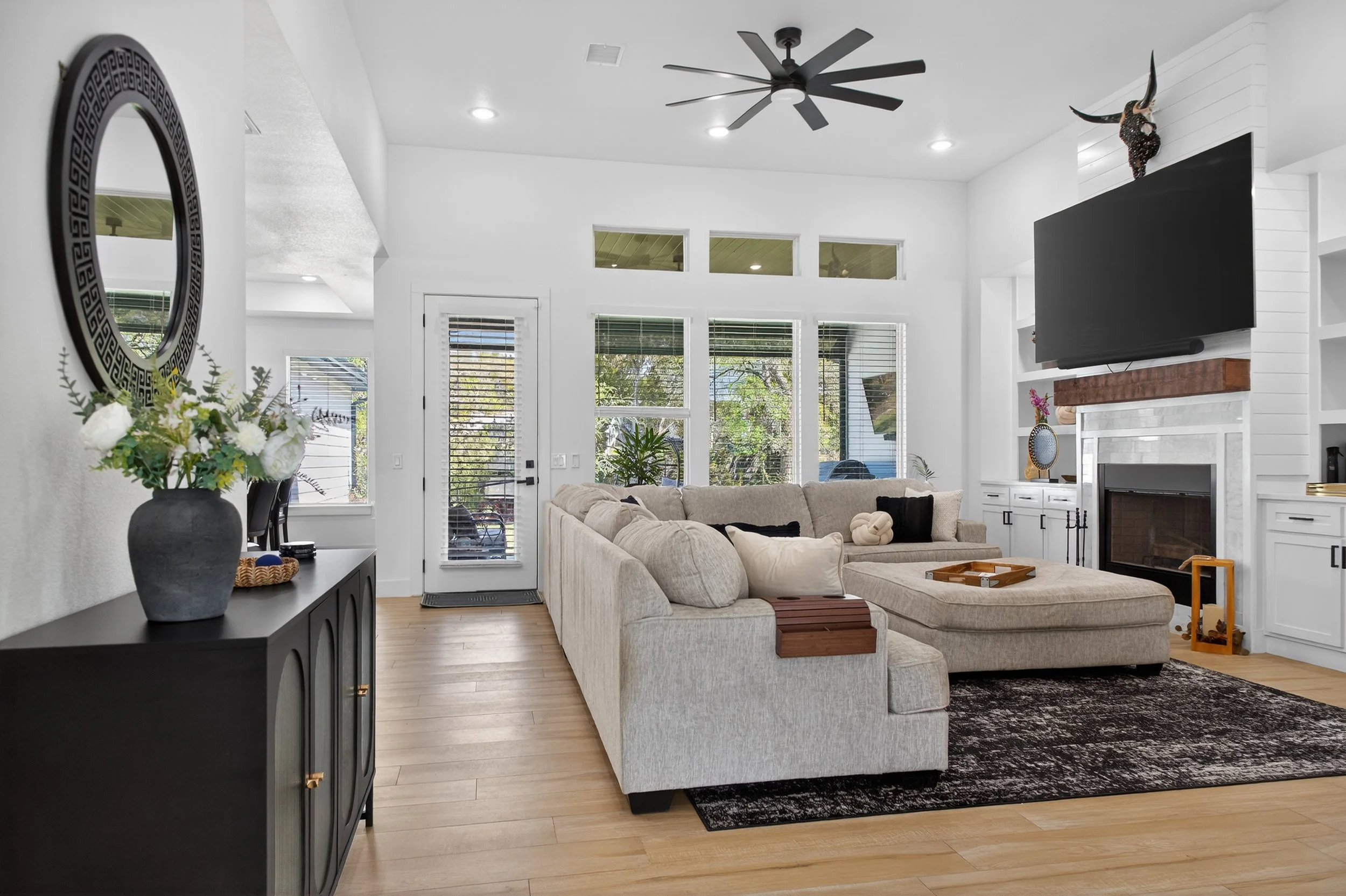 A bright living room with white walls, large windows, a beige sectional sofa, a black sideboard with a flower vase, a wall-mounted flat-screen TV, and decorative items including a mounted animal skull with horns.