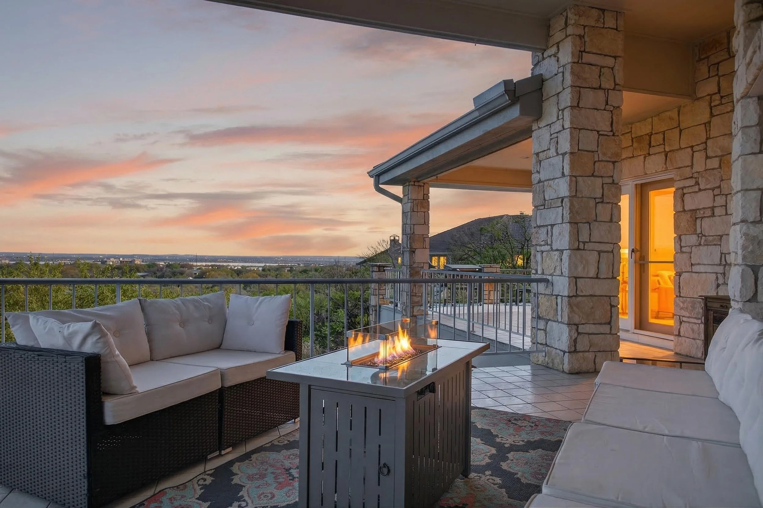 A cozy outdoor patio with white cushioned seating, a firepit table, and a stone building with a glowing interior door, overlooking a scenic view of a sunset sky and distant trees and houses.